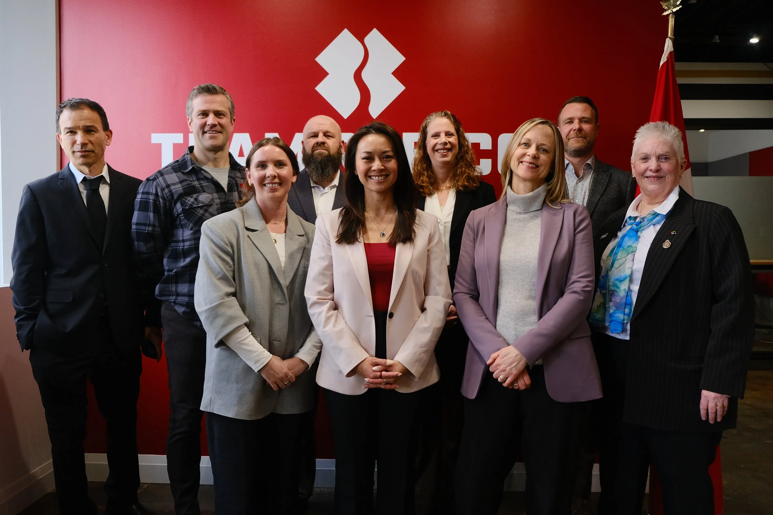 A group of nine diverse professionals posing for a photo in front of a red TAMA logo background.