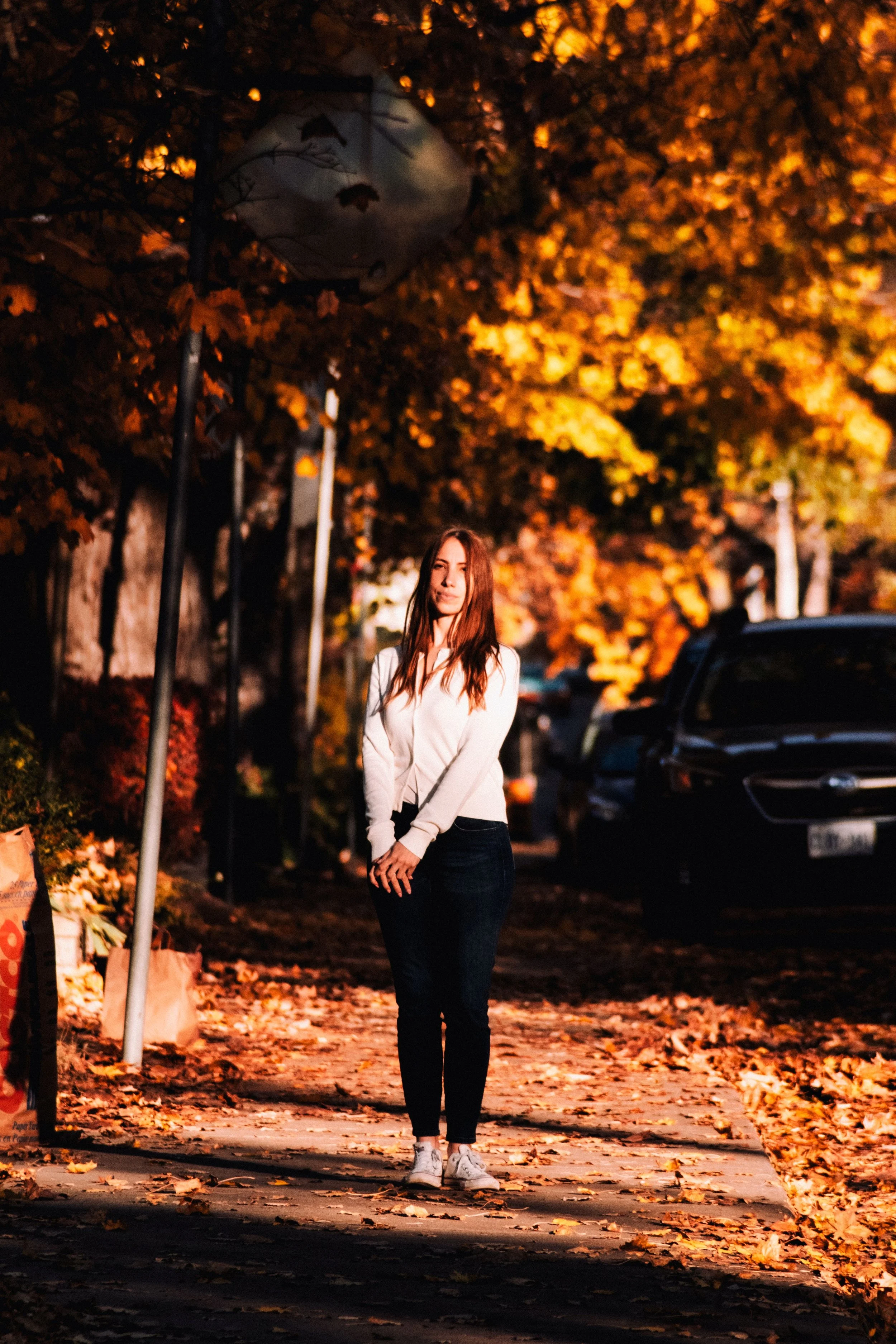 A woman stands on a sidewalk covered with fallen autumn leaves, facing the camera, surrounded by orange and yellow trees, with parked cars in the background during late afternoon or early evening.