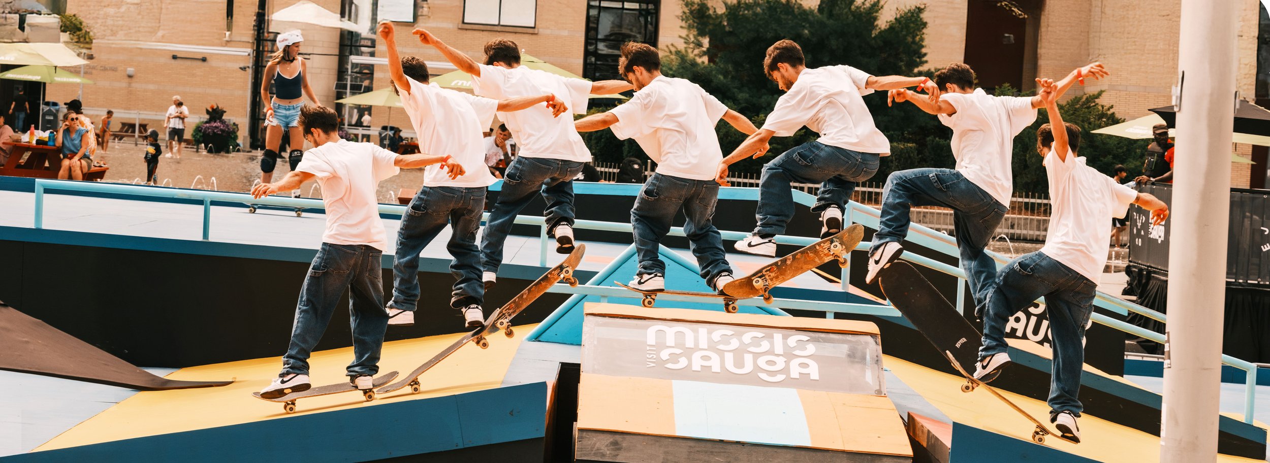 Multiple boys skateboarding at a skate park, performing tricks on a ramp with a sign that says "Visit Mission Sauga" in the foreground. Bright, sunny day with onlookers in the background.