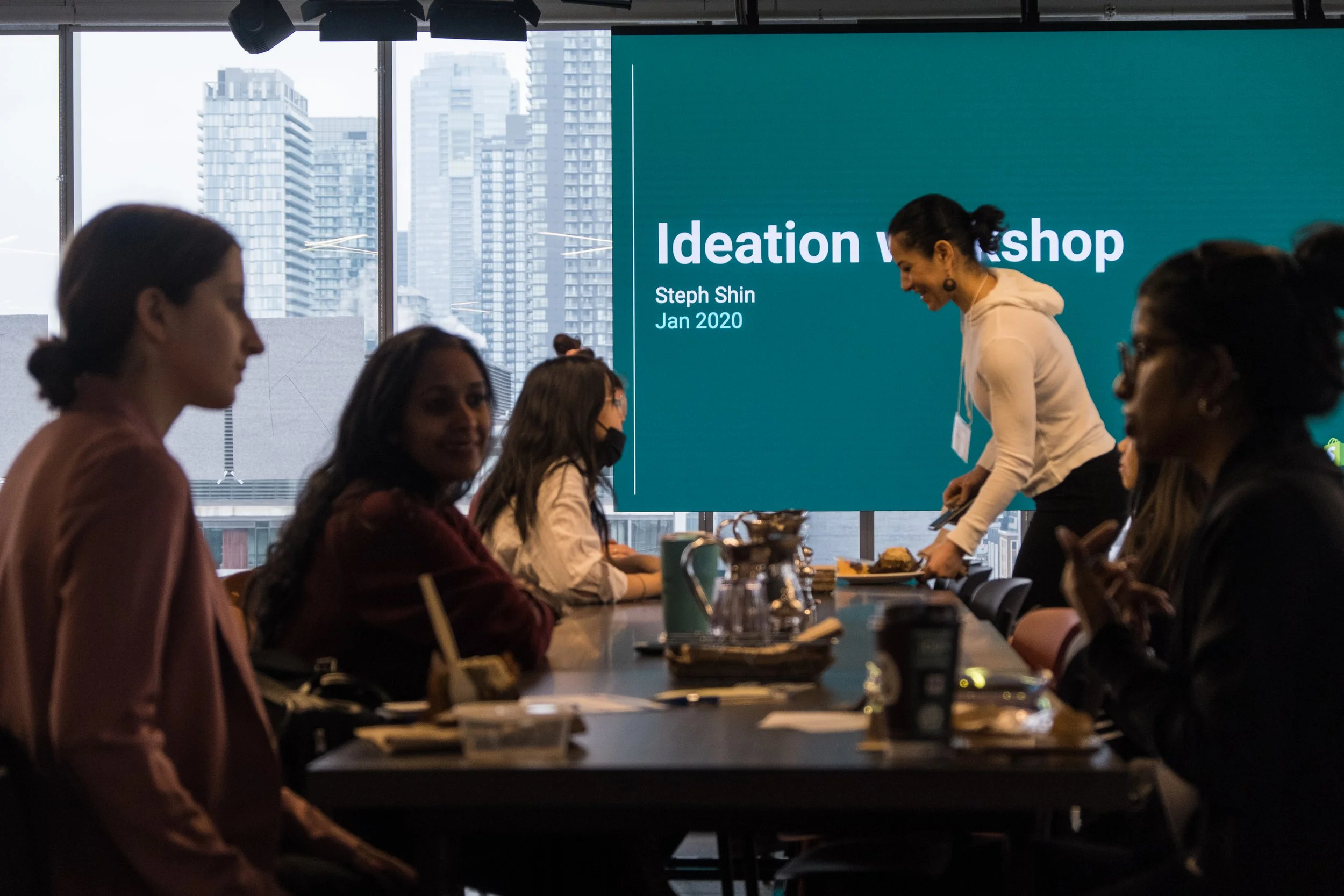 A group of women attending a workshop in a conference room with large windows and a city skyline. A woman is serving food, and a large screen displays 'Ideation Workshop, Steph Shin, Jan 2020'.