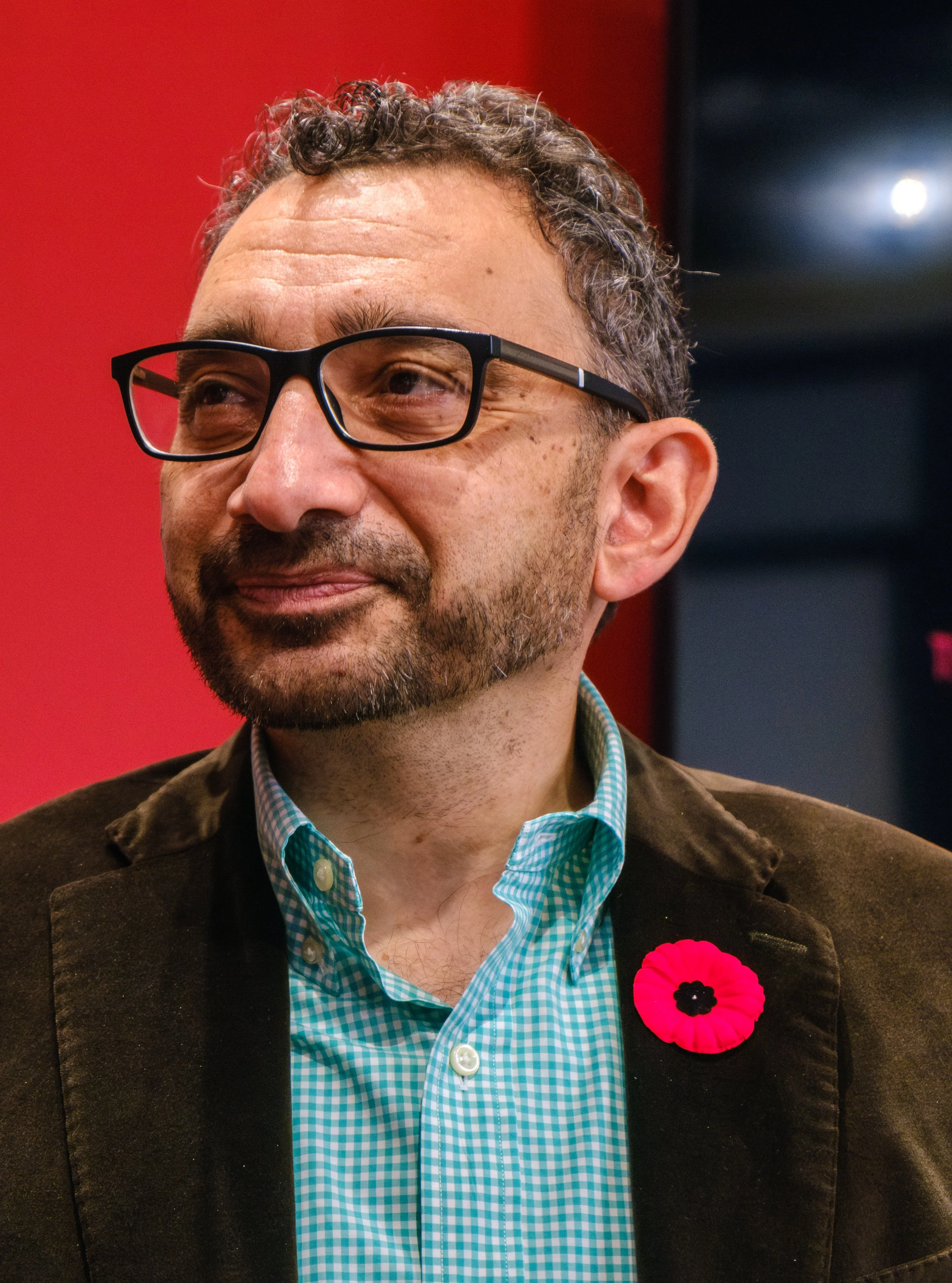 A man wearing glasses, a blue checkered shirt, and a brown jacket, with a pink poppy pin on his jacket lapel, standing in front of a red background.