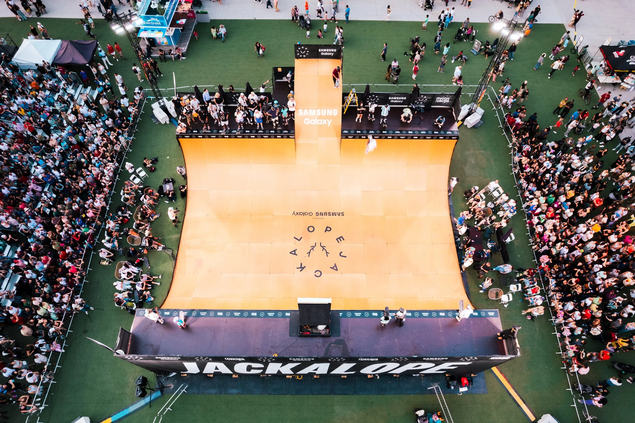 Overhead view of a large crowd watching a skateboarding event at a skate rink with a Jump Attack sign, a Samsung Galaxy branding, and a ramp with a yellow surface for skateboarding.