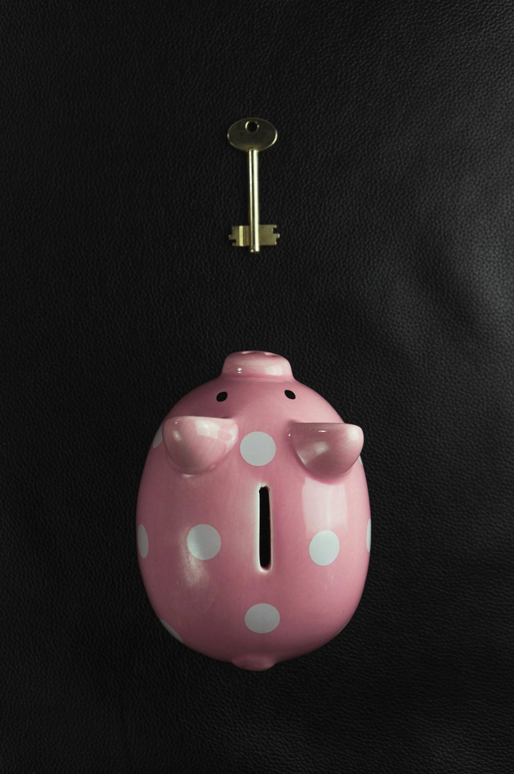 Pink polka dot piggy bank with a coin slot on its back, positioned on a black surface, with a key hanging above on a black background.