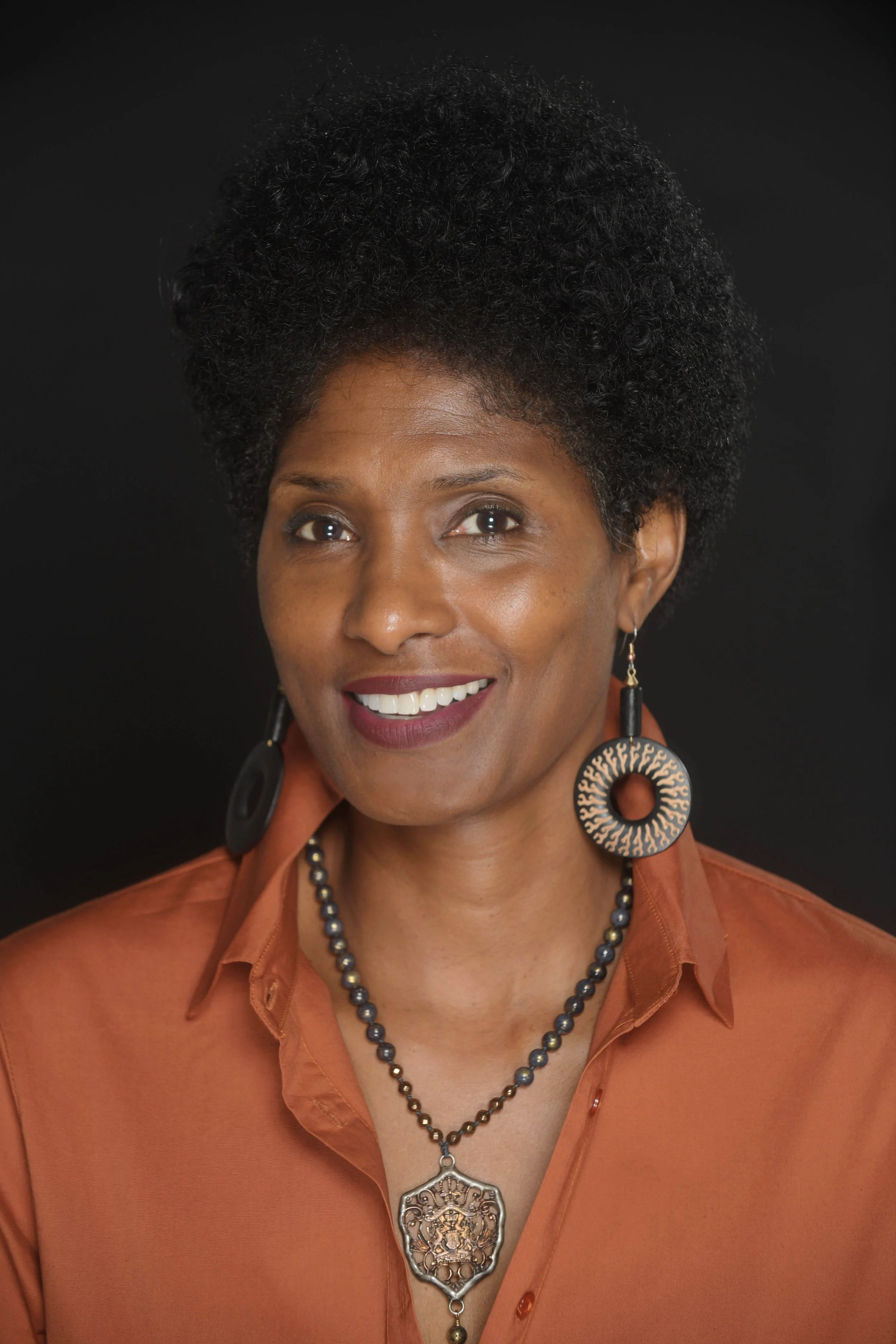 A woman with short, curly black hair, wearing large circular earrings with patterns, a beaded necklace, and an orange shirt, smiling against a black background.
