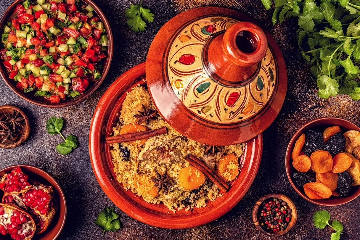 A traditional Moroccan tagine dish with couscous, vegetables, and spices, served in an earthenware tagine pot, surrounded by bowls of salad, dried fruits, and spices, with fresh herbs for garnish.