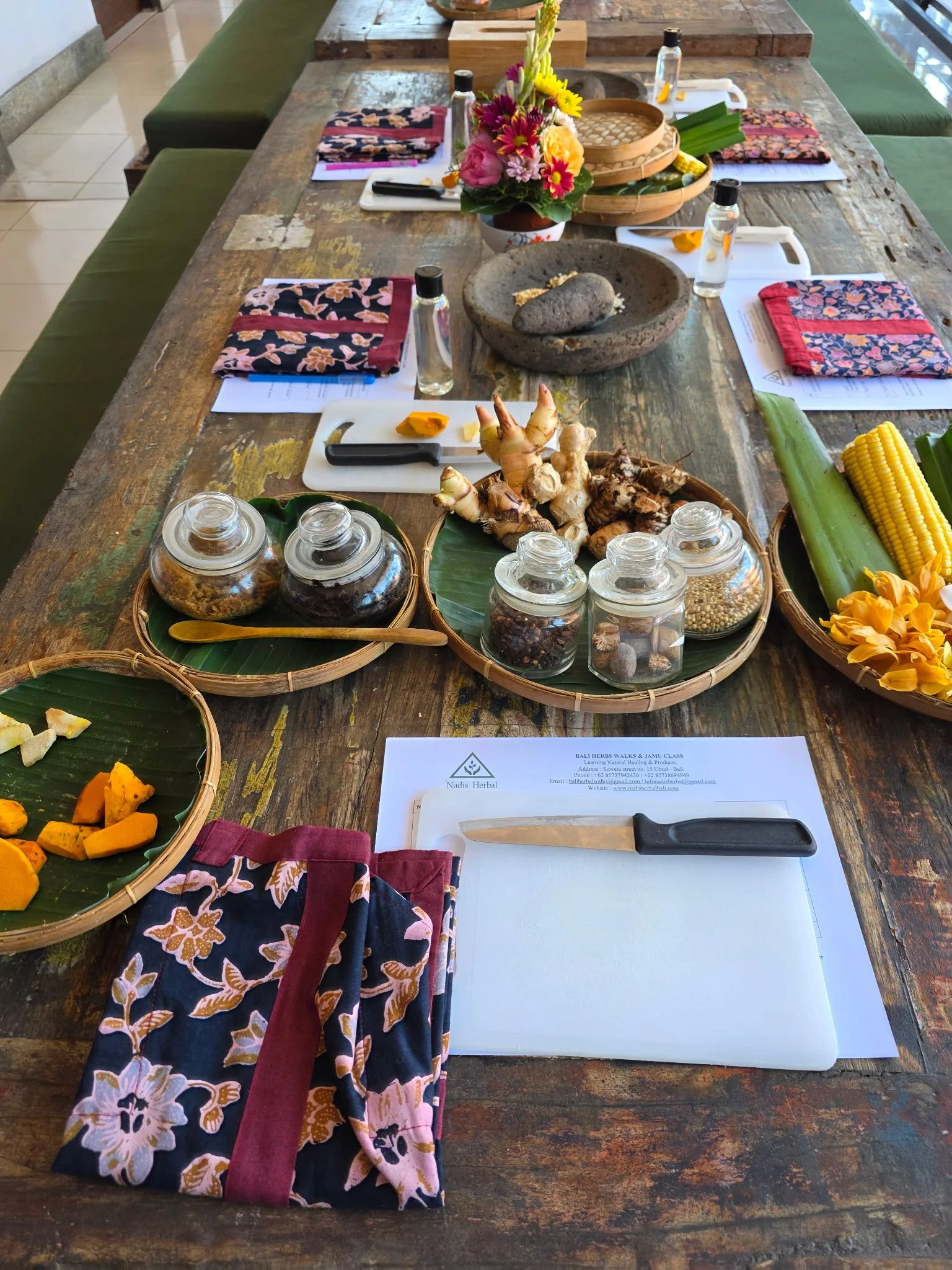 A rustic wooden table set up for a herbal medicine or wellness workshop with various herbs, spices, and ingredients in jars, along with flowers, snacks, and a knife on a white cutting board.
