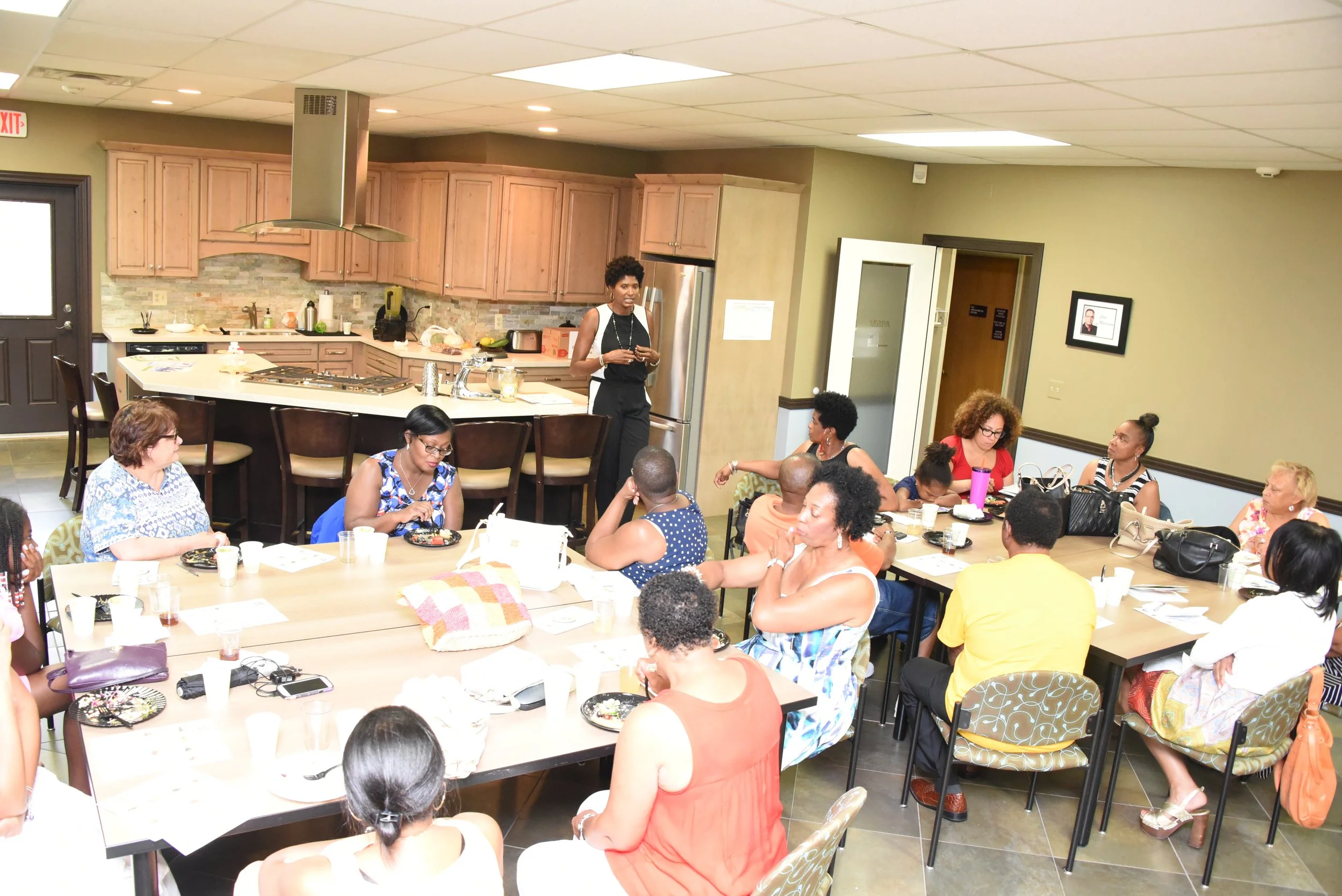 A group of women and a few children are sitting around tables in a room with a kitchen in the background. A woman is standing and speaking to the group.
