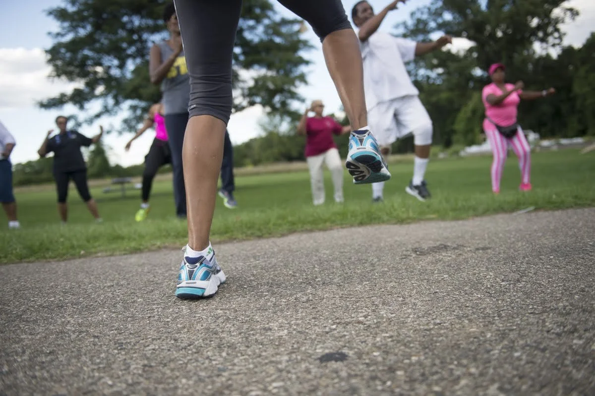 A group of people exercising outdoors in a park, with a focus on a woman in running shoes performing jumping exercises in the foreground. Other participants are visible in the background, doing similar movements.