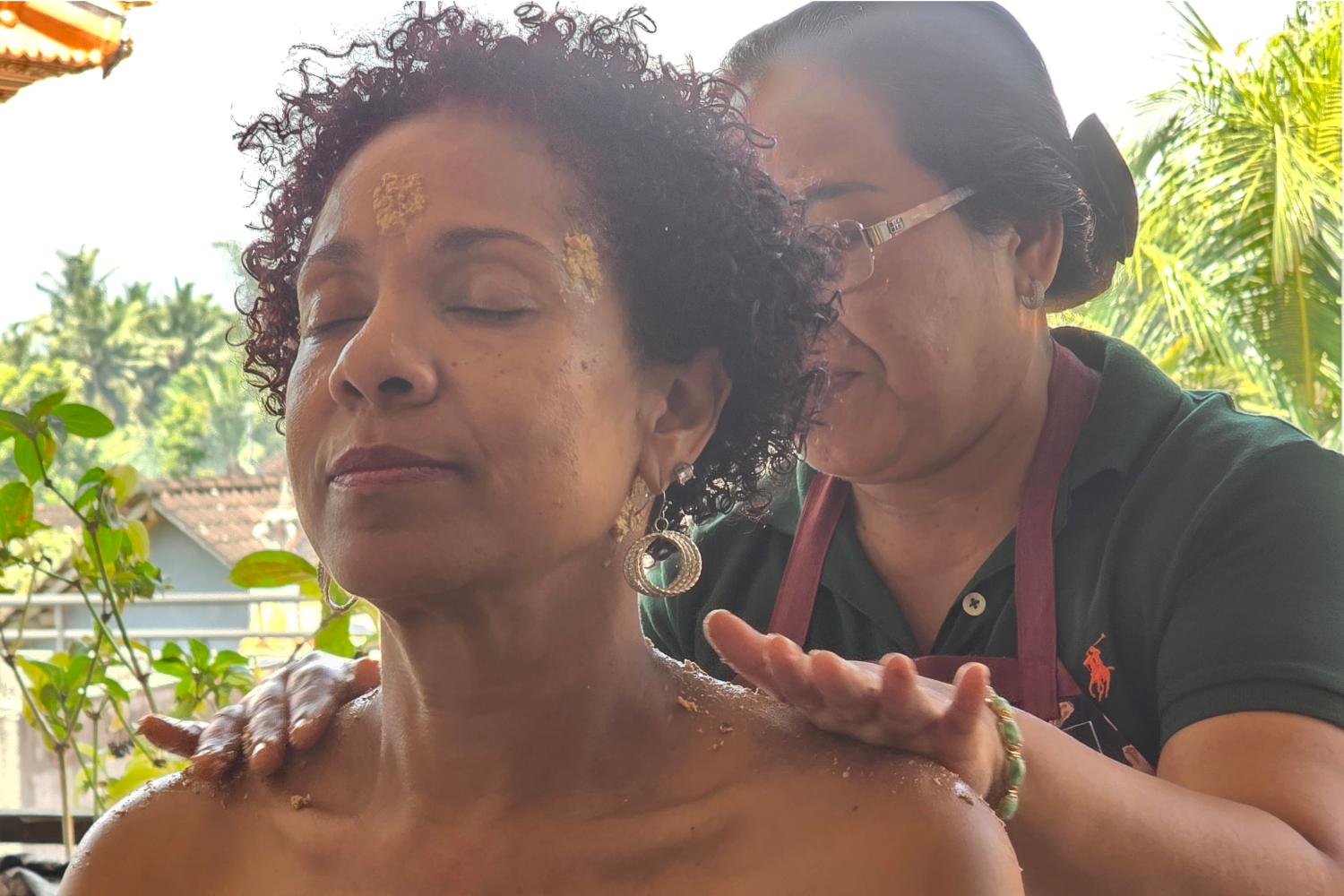 A woman with closed eyes receiving a massage outdoors, with a massage therapist applying oil on her shoulder, surrounded by green plants and trees.
