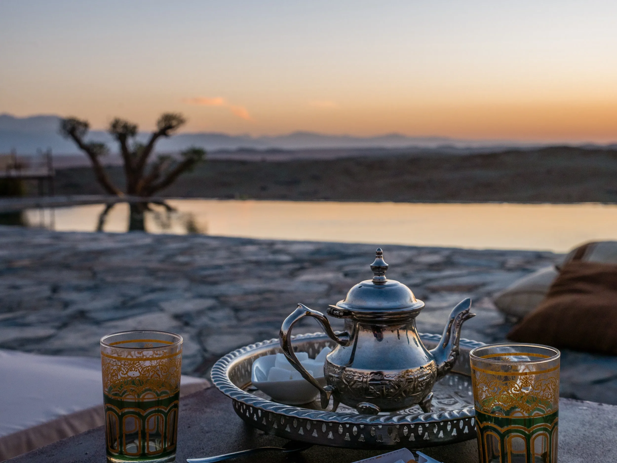 Tea set with two glasses on a tray, outdoor desert landscape at sunset with a tree reflection in water in the background.
