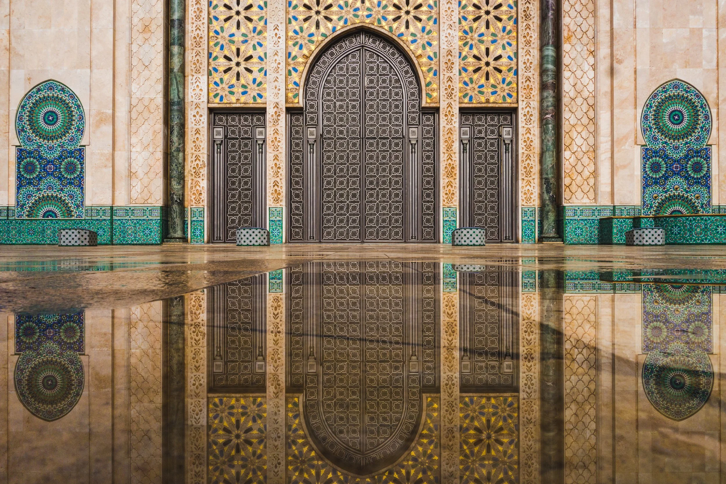 Intricate Moroccan-style architecture with ornate black door, blue and green mosaic tiles, and reflection on polished floor.