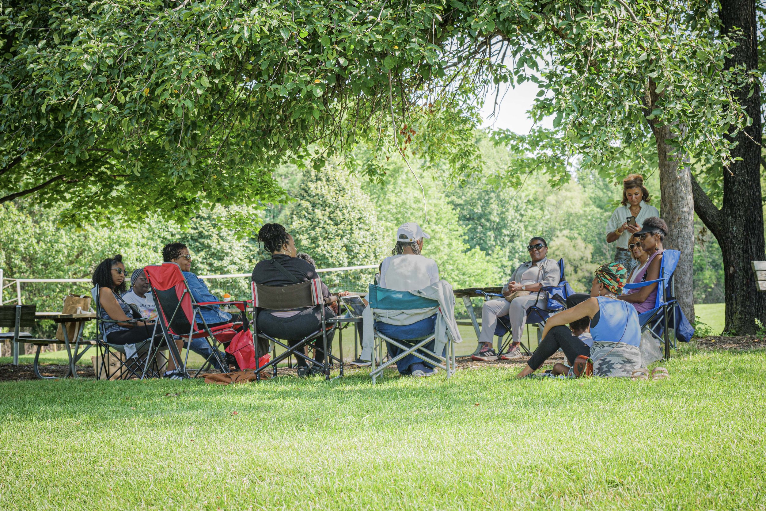 A group of women seated in a circle on lawn chairs under trees in a park, engaging in conversation.