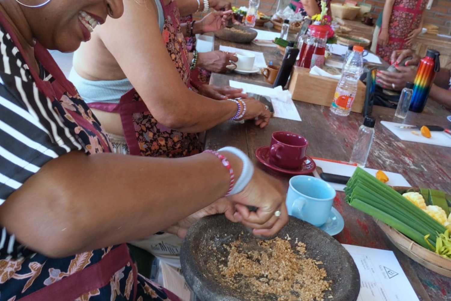 People gathered around a wooden table participating in a food preparation activity, with various cups, bottles, and food items on the table, and one person grinding ingredients in a stone mortar.