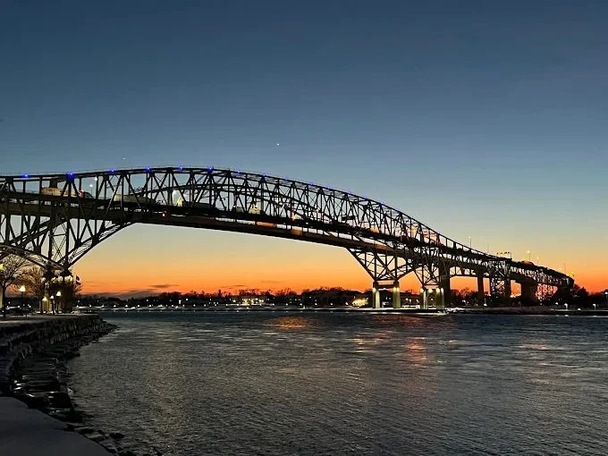 A large bridge over a body of water at sunset, with a clear sky and city lights in the distance.