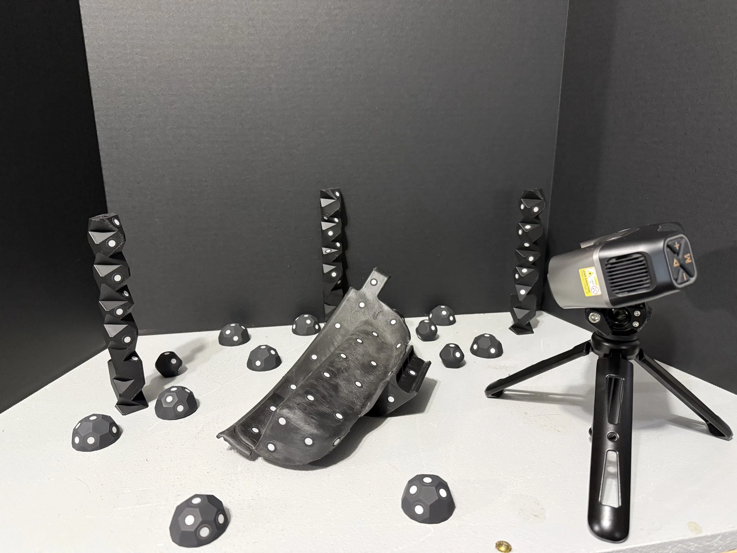 A tabletop with black and gray geometric objects resembling dice, some on a black stand, and a mounted laser measuring device on a tripod.