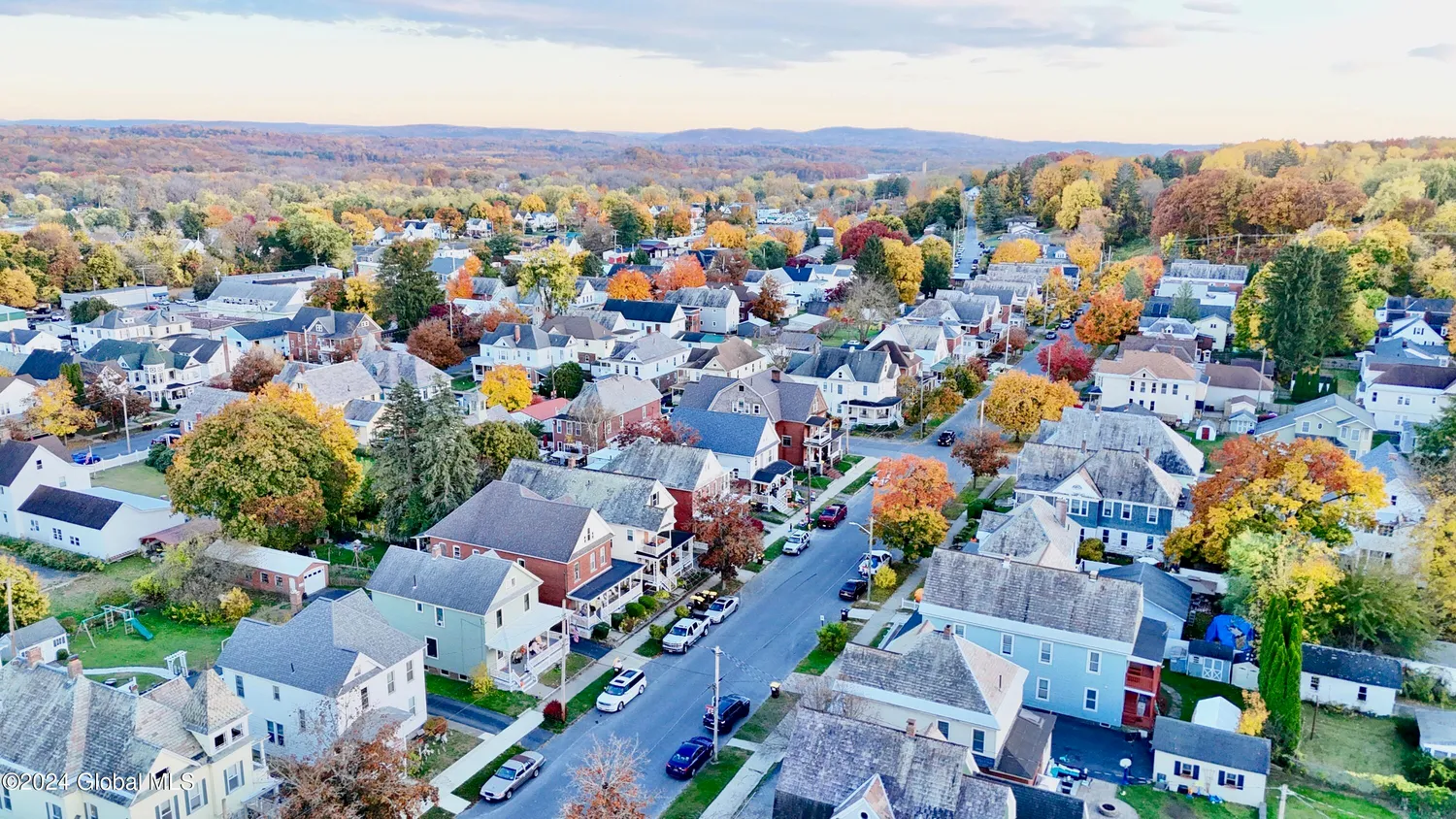 An aerial view of a suburban neighborhood with numerous houses, trees with fall foliage, and a street lined with parked cars, with a hilly landscape in the background.