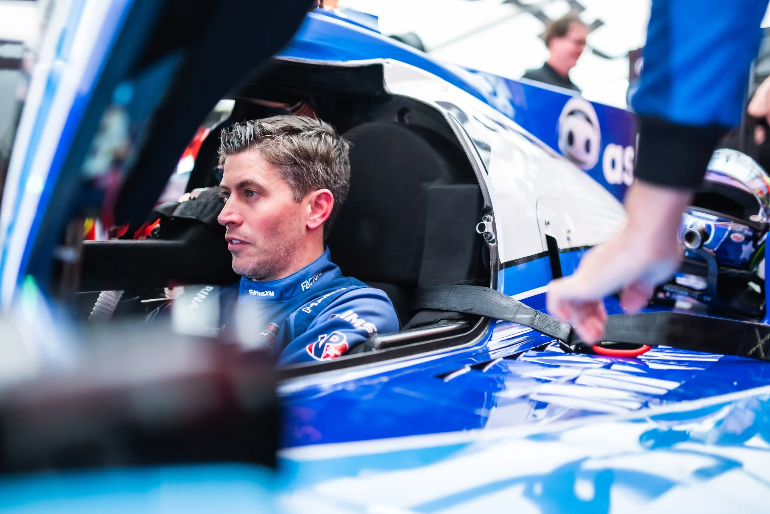 A race car driver sitting inside a blue racing car, preparing for a race, with a team member adjusting equipment nearby.