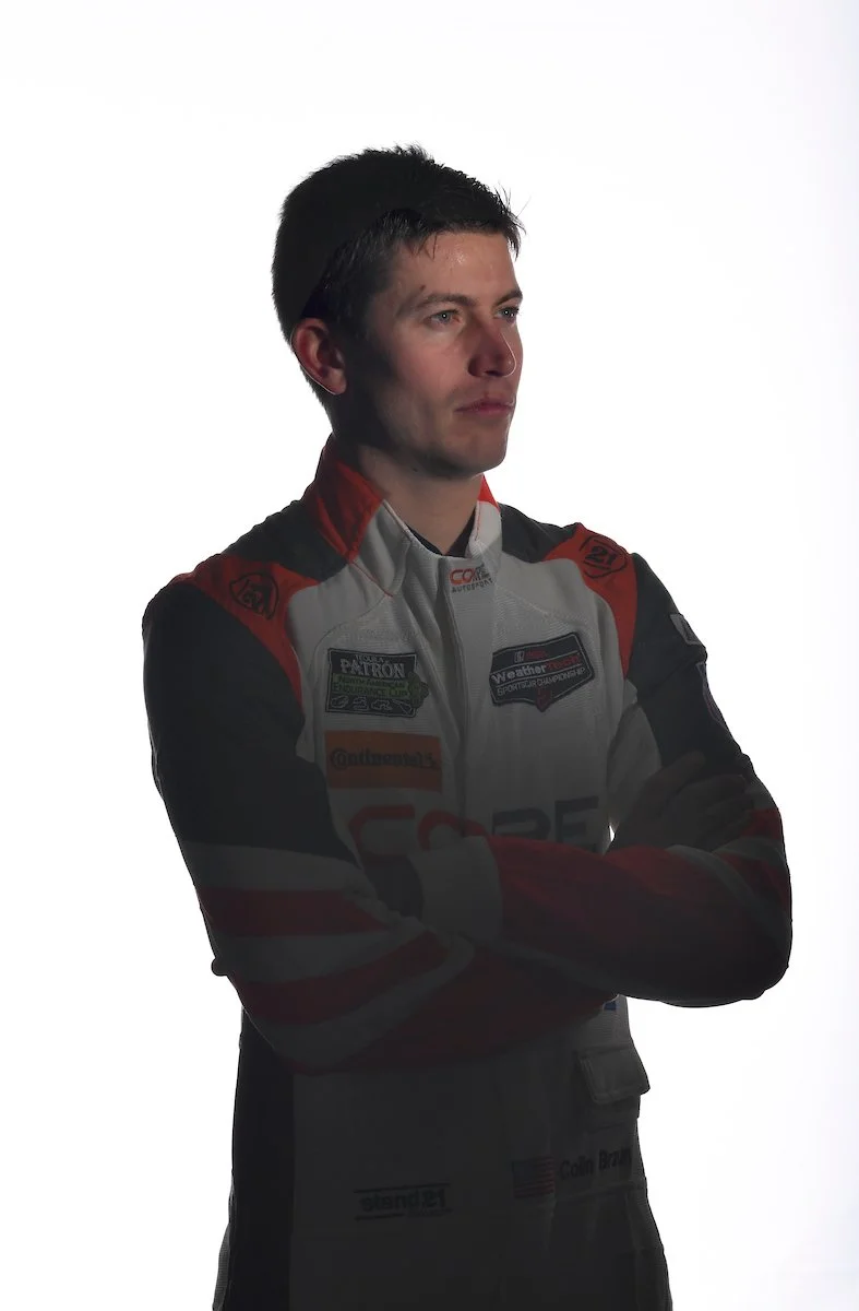 A male race car driver in a racing suit with arms crossed, facing slightly to the right, against a plain white background.