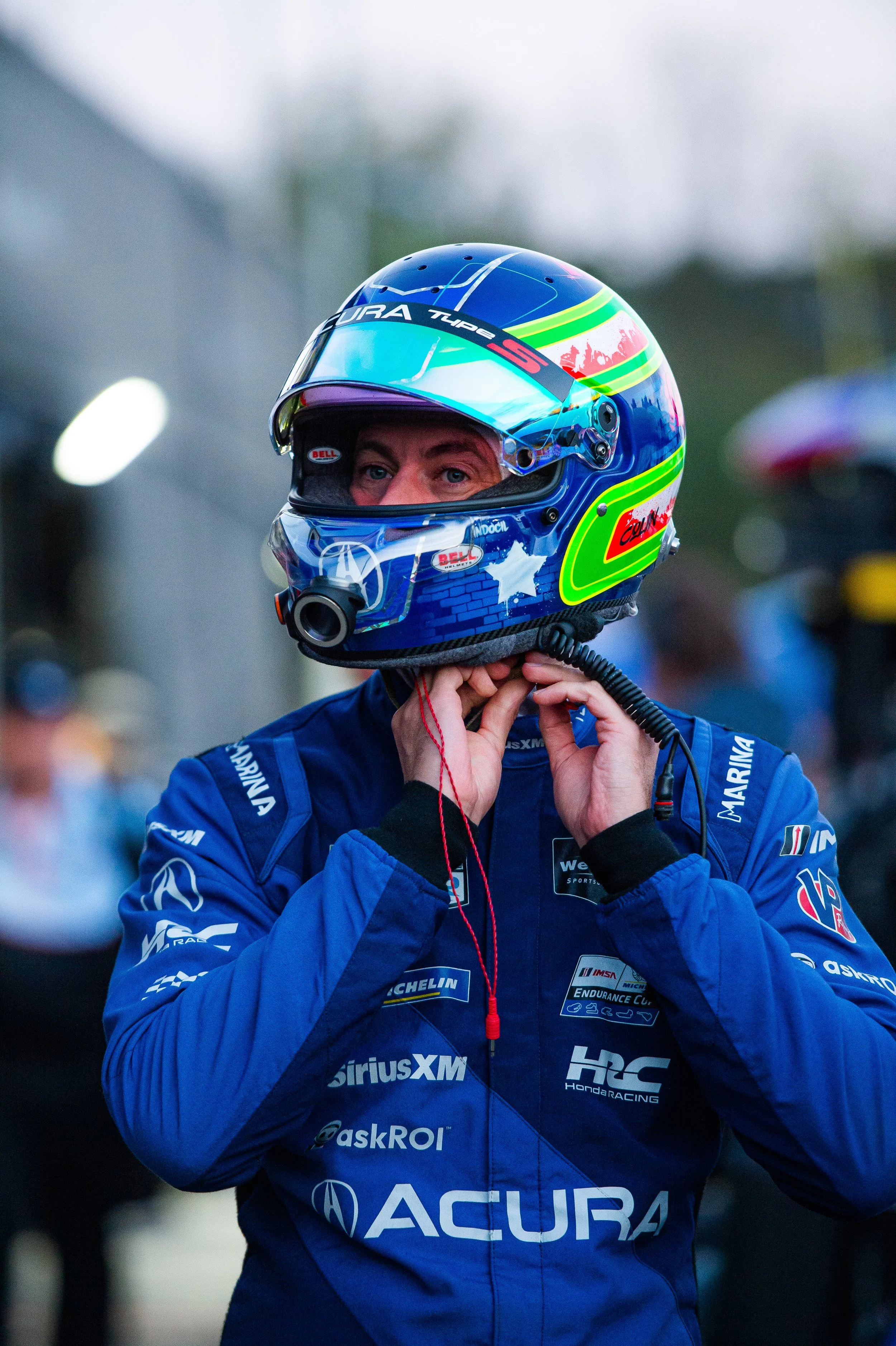 A race car driver in a blue racing suit putting on a multicolored helmet with reflective visor, standing in a racing paddock area.