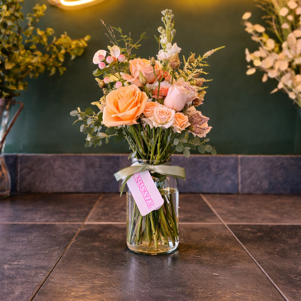 A bouquet of peach and white roses, pink berries, and greenery in a clear glass jar with a green ribbon and a pink price tag, placed on a dark tiled surface.