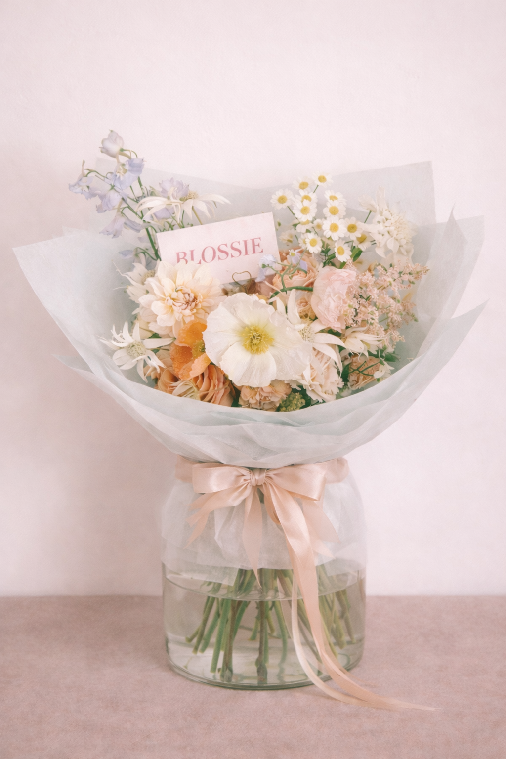 A bouquet of pastel-colored flowers in a glass vase with a pink satin ribbon, including daisies, roses, and other blooms, with a card that says 'BLOSSIE'.