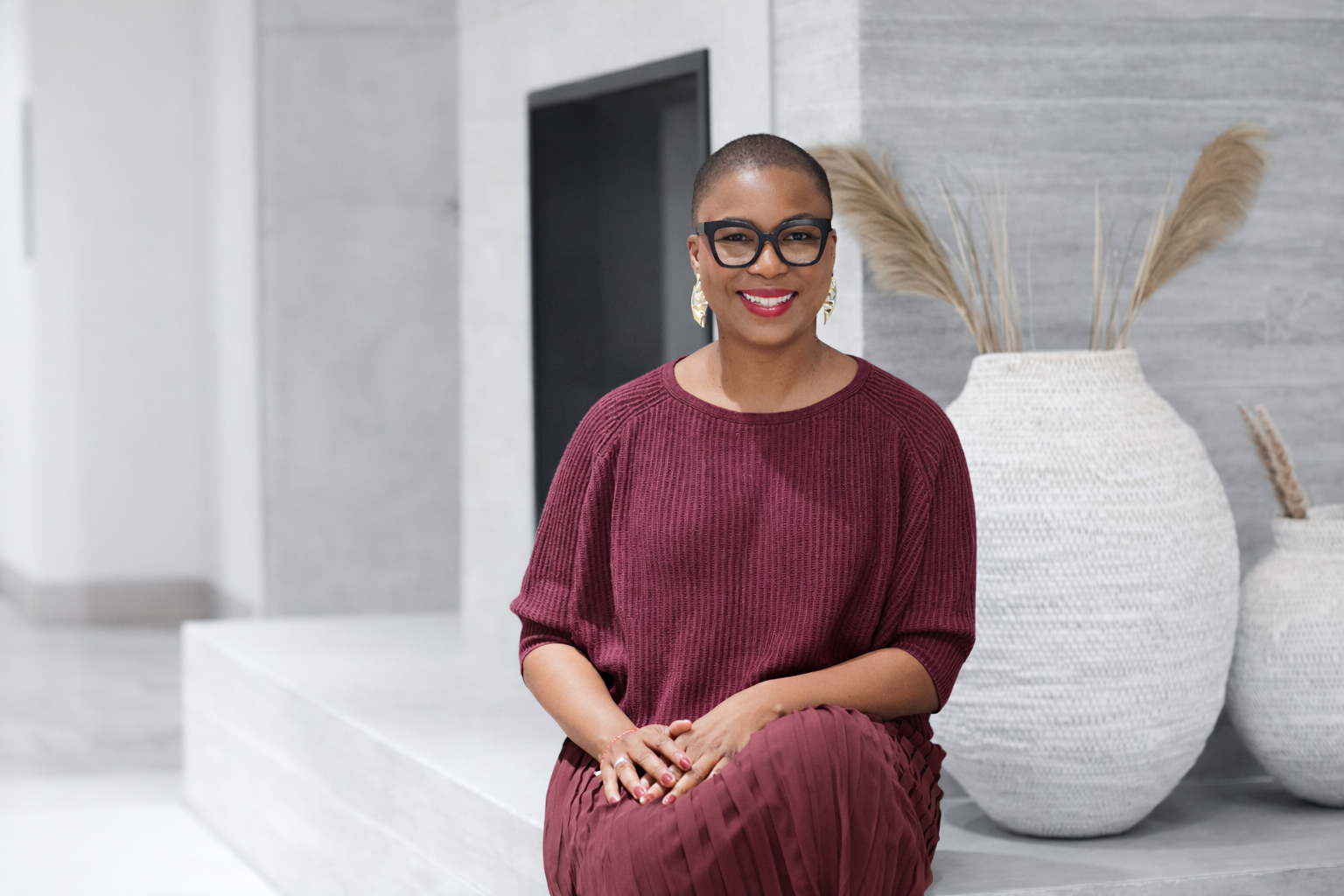 A woman with short hair, glasses, and gold earrings, wearing a burgundy sweater and skirt, sitting on a white platform with large white vases and dried plants in the background.