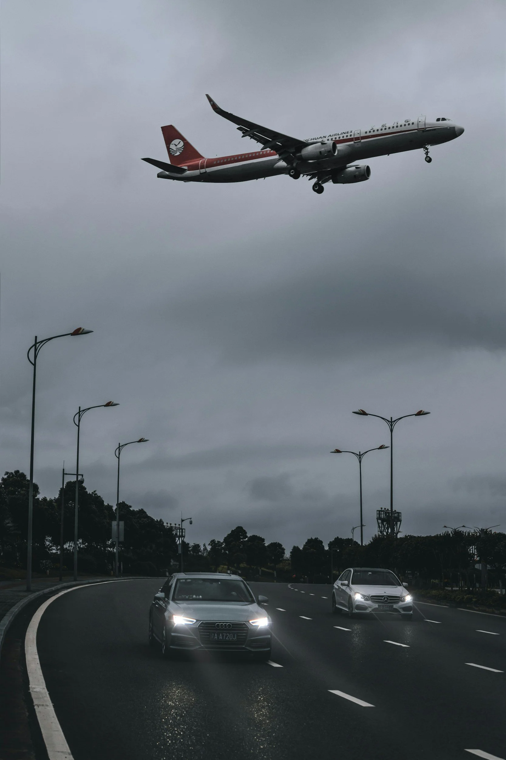 An airplane flying low over a curved city road with cars, trees, and street lamps under a cloudy sky.