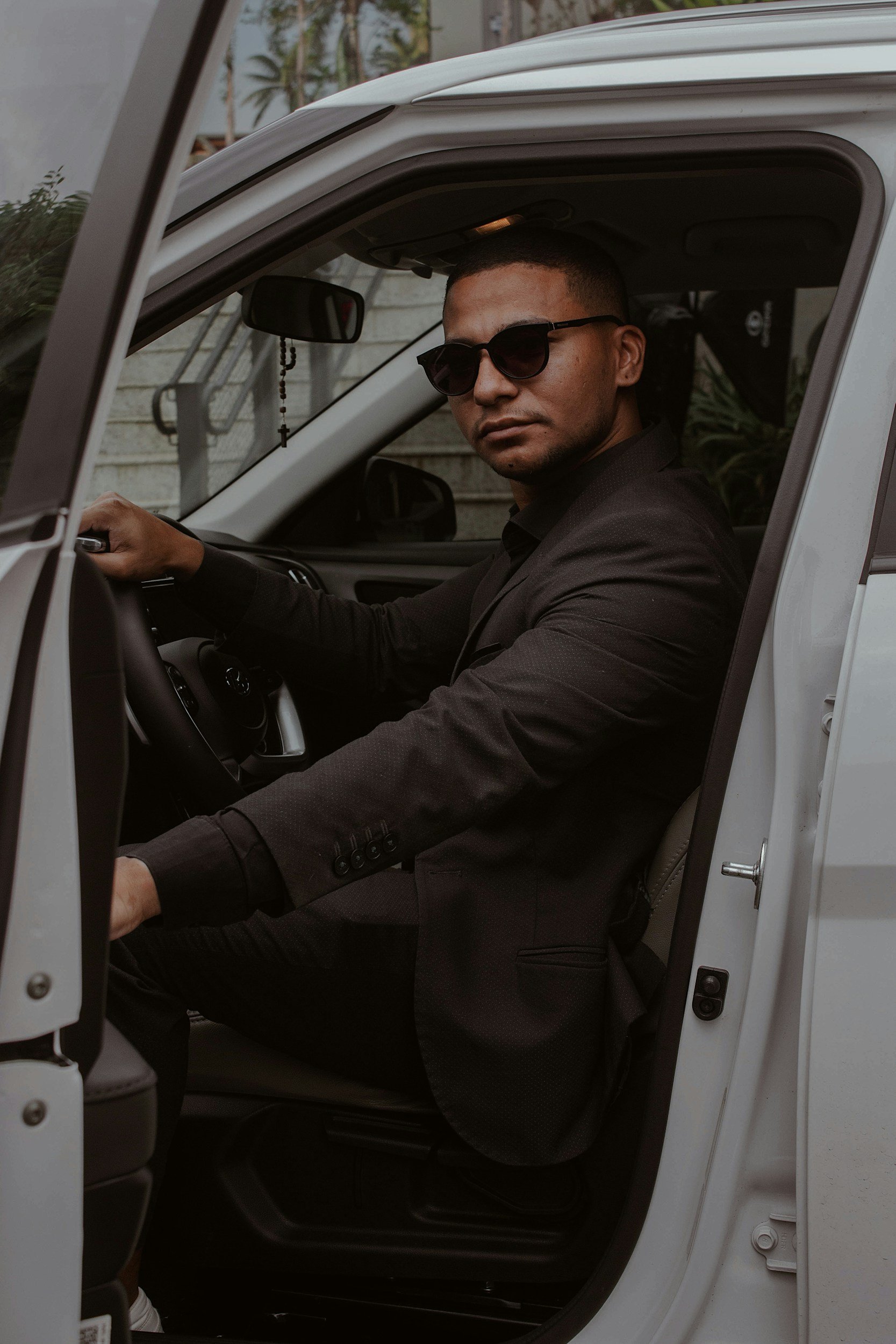 A man wearing sunglasses and a black suit sitting in the driver's seat of a white car.