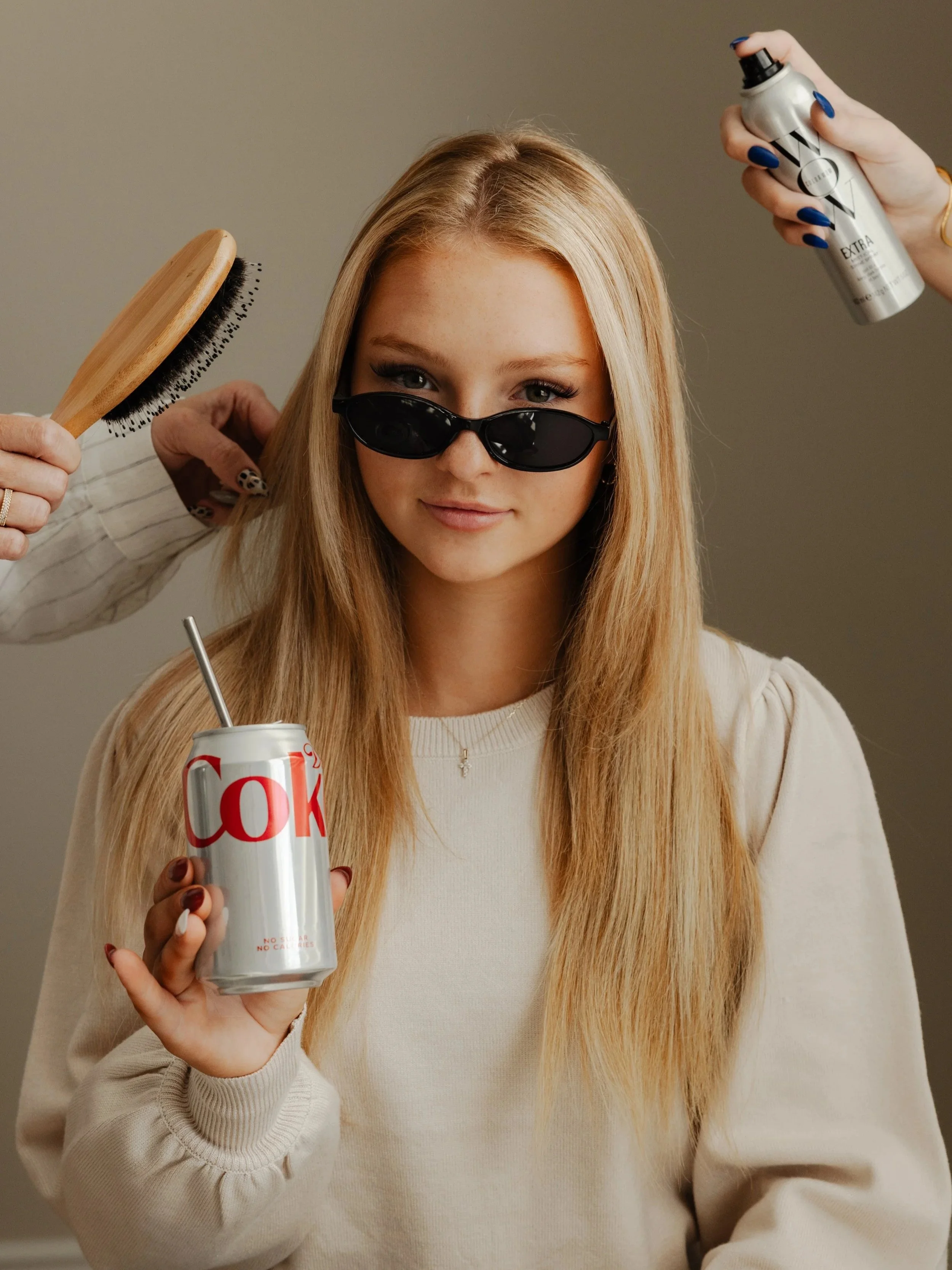 A young woman with long blonde hair wearing black sunglasses and a cream-colored sweater, holding a can of Diet Coke with a straw in her right hand, while hair styling tools and products are positioned around her head.
