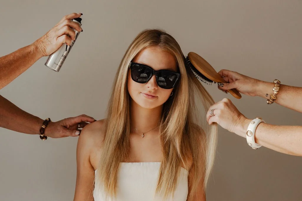 A woman with long blonde hair wearing black sunglasses and a white strapless top while getting her hair styled by multiple people, with one person using a spray can, another brushing her hair, and others touching her hair.