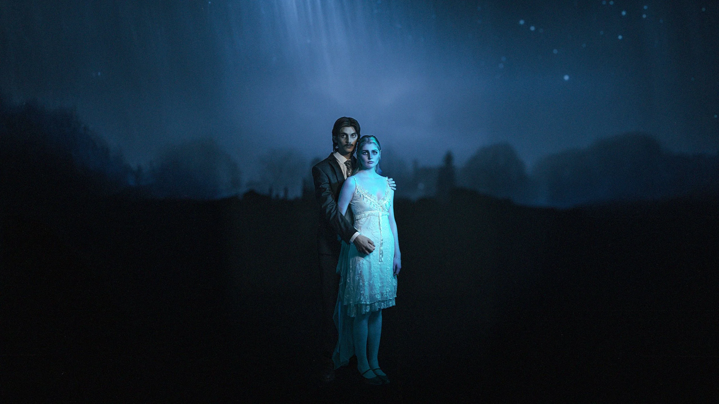 En mann og en kvinne står sammen utendørs om natten, med mørkt landskap og stjernehimmel bak seg. Mannen er iført dress, og kvinnen har på seg en blondet kjole, opplyst av blålig lys.