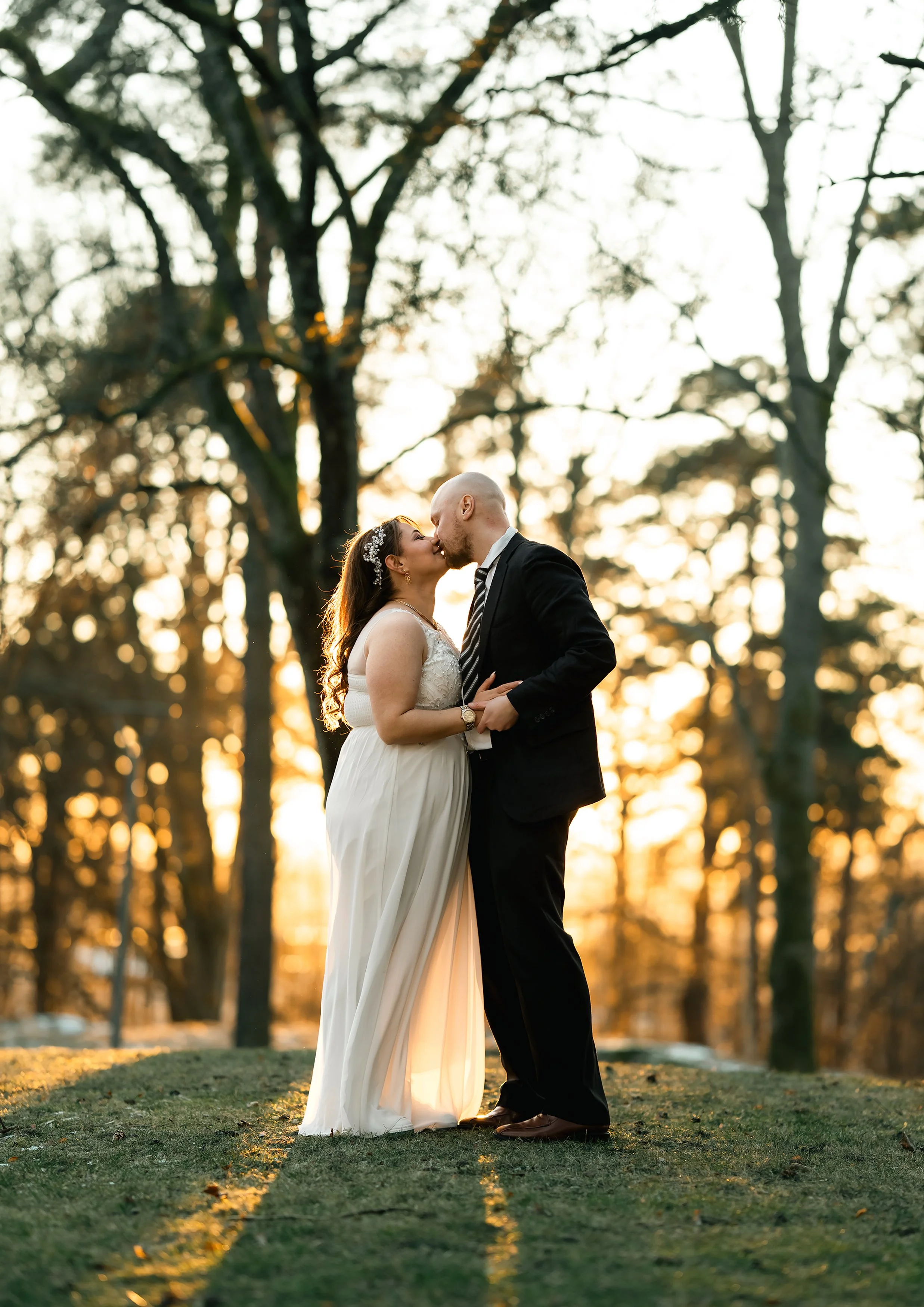 Et brudepar i bryllupsantrekk kysser hverandre utendørs i solnedgang, med trær i bakgrunnen.