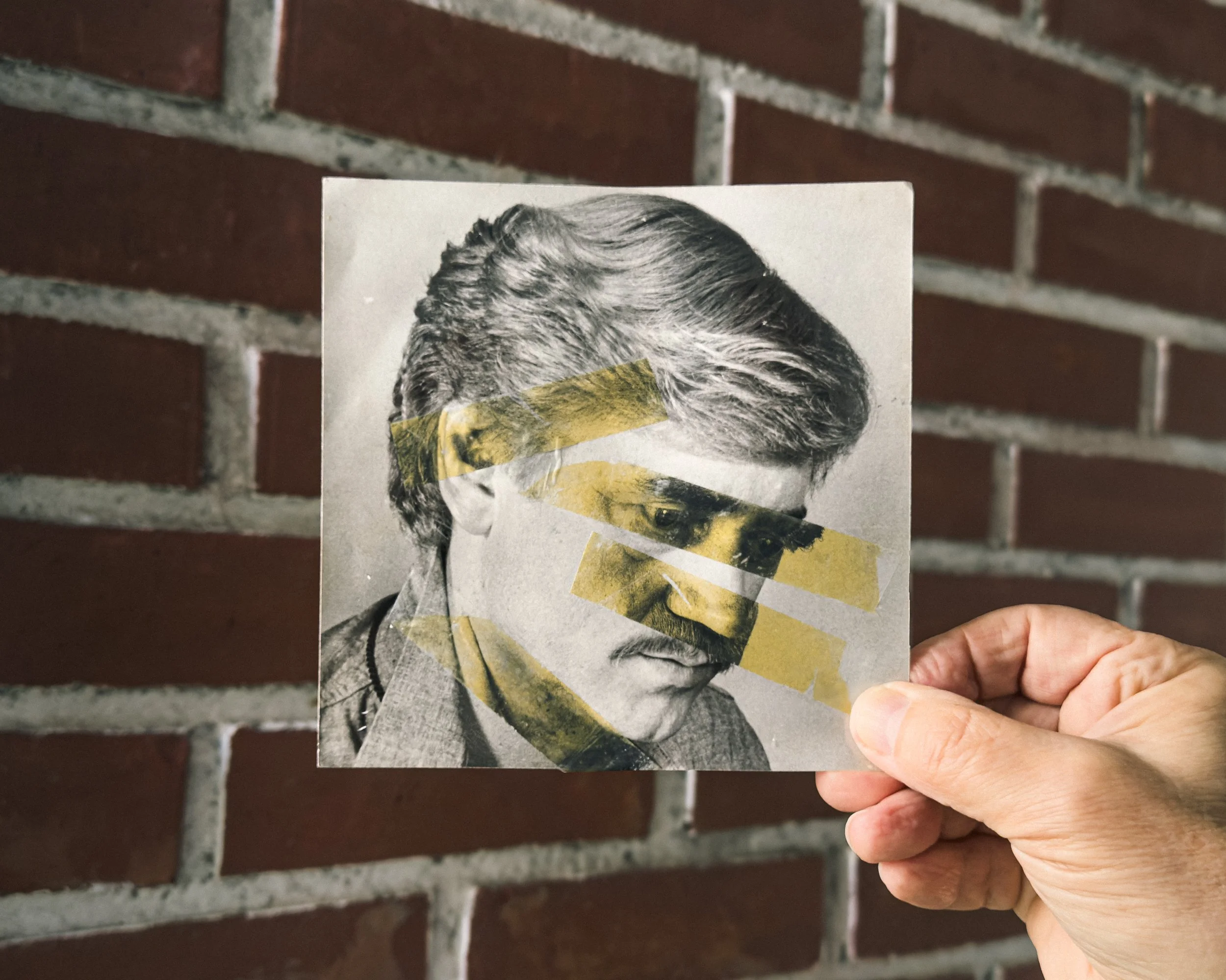 En hånd holder et svart-hvitt-fotografi av en mann med stylet hår og bart, dekket med gule tape-markeringer, mot en bakgrunn av rød murvegg.