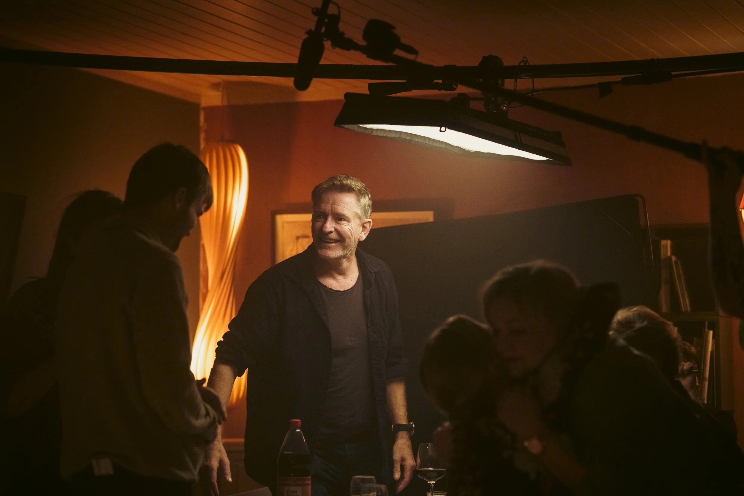 En scene fra et film- eller TV-opptak der en smilende mann er omgitt av crew og teknisk utstyr, badet i varmt lys