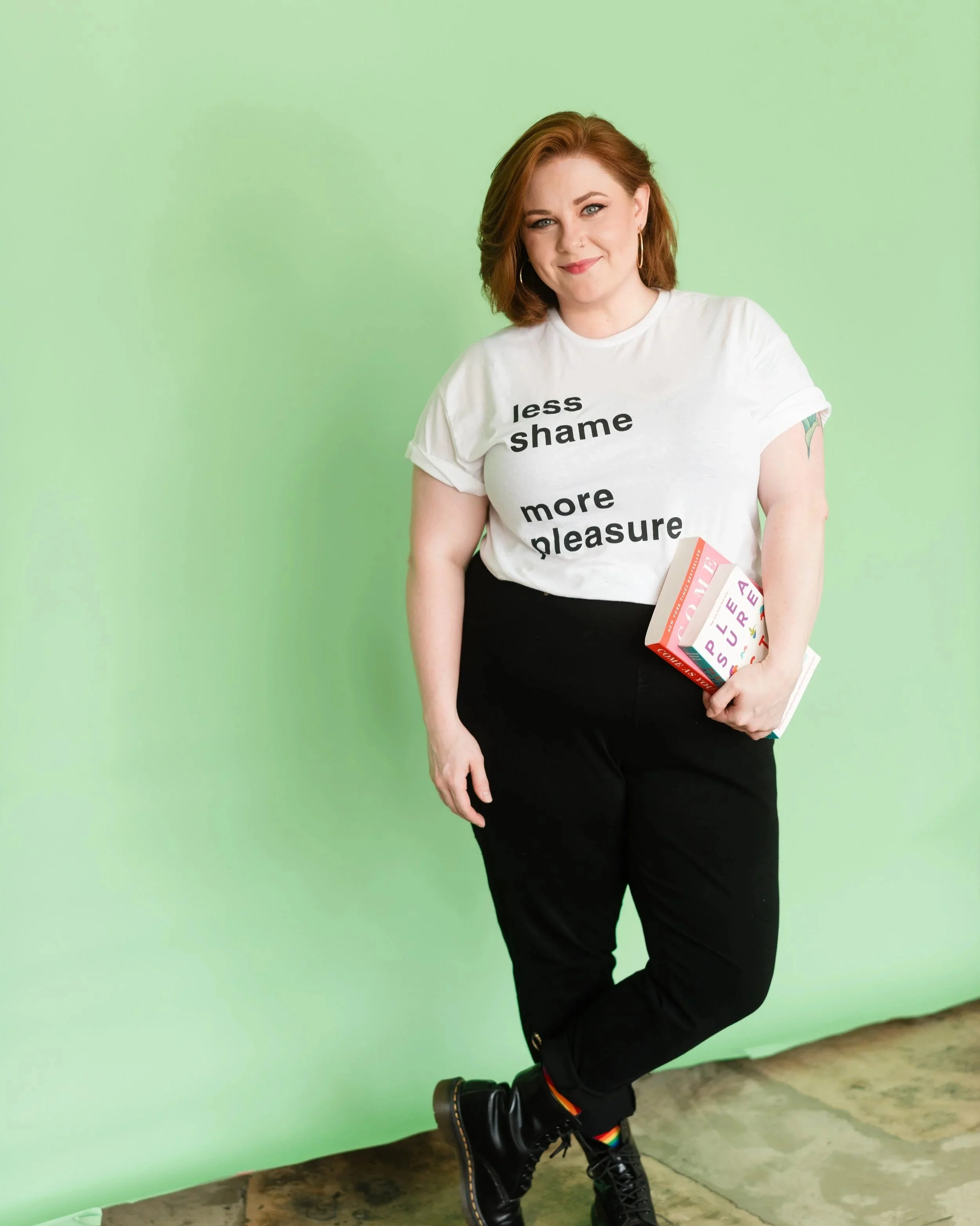 A woman with red hair, wearing a white t-shirt with black text, black pants, and black boots with rainbow laces, standing against a green wall, holding books.