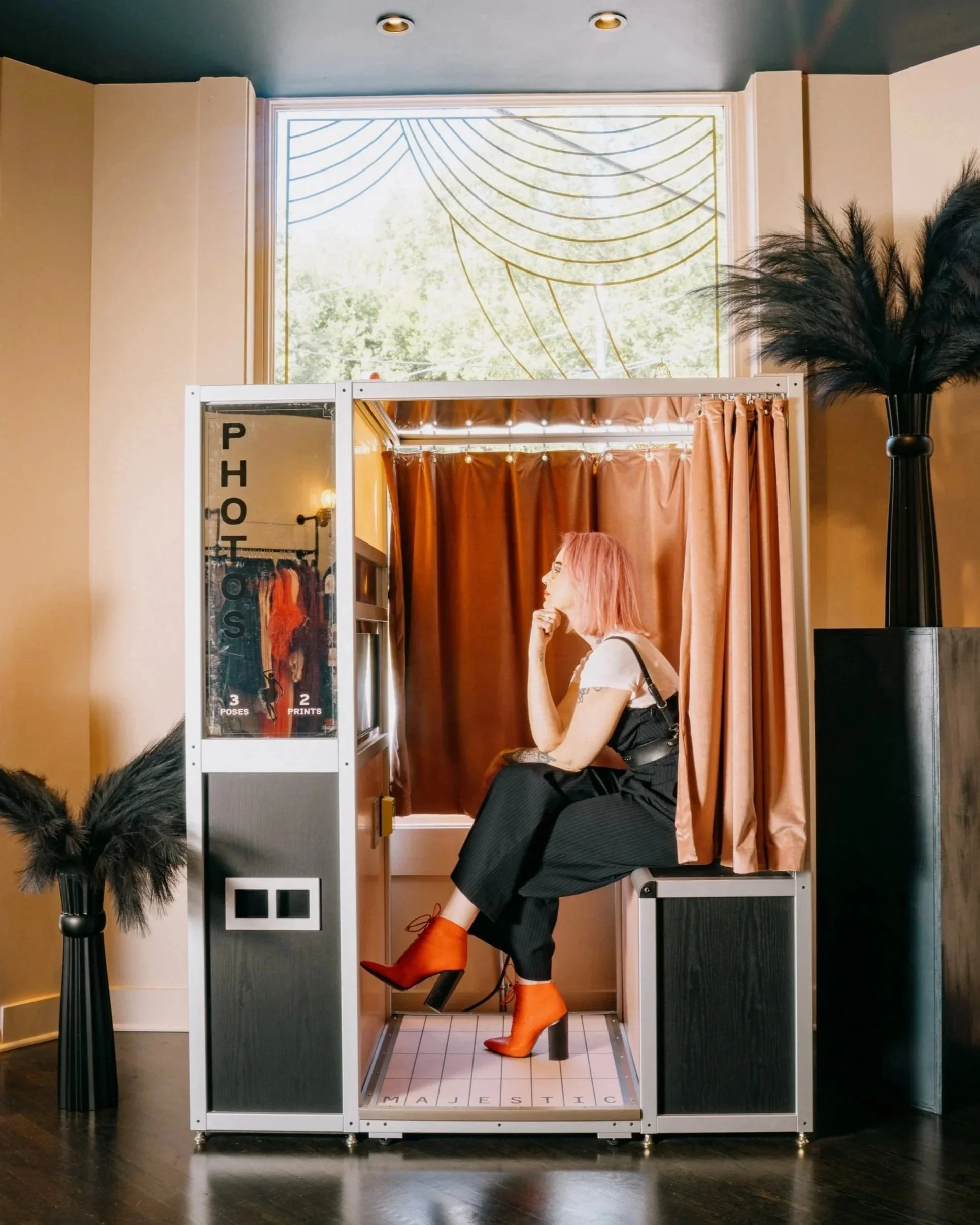 A woman with pink hair sitting inside a photo booth with orange boots, black pants, white shirt, and black vest. The photo booth has a white frame, brown curtains, and a digital mirror with pose and print options. The booth is on a tile floor with 'MAJESTIC' spelled out at the bottom, next to a black vase with black feathers. A large window with decorative gold lines and green trees outside is above the booth.