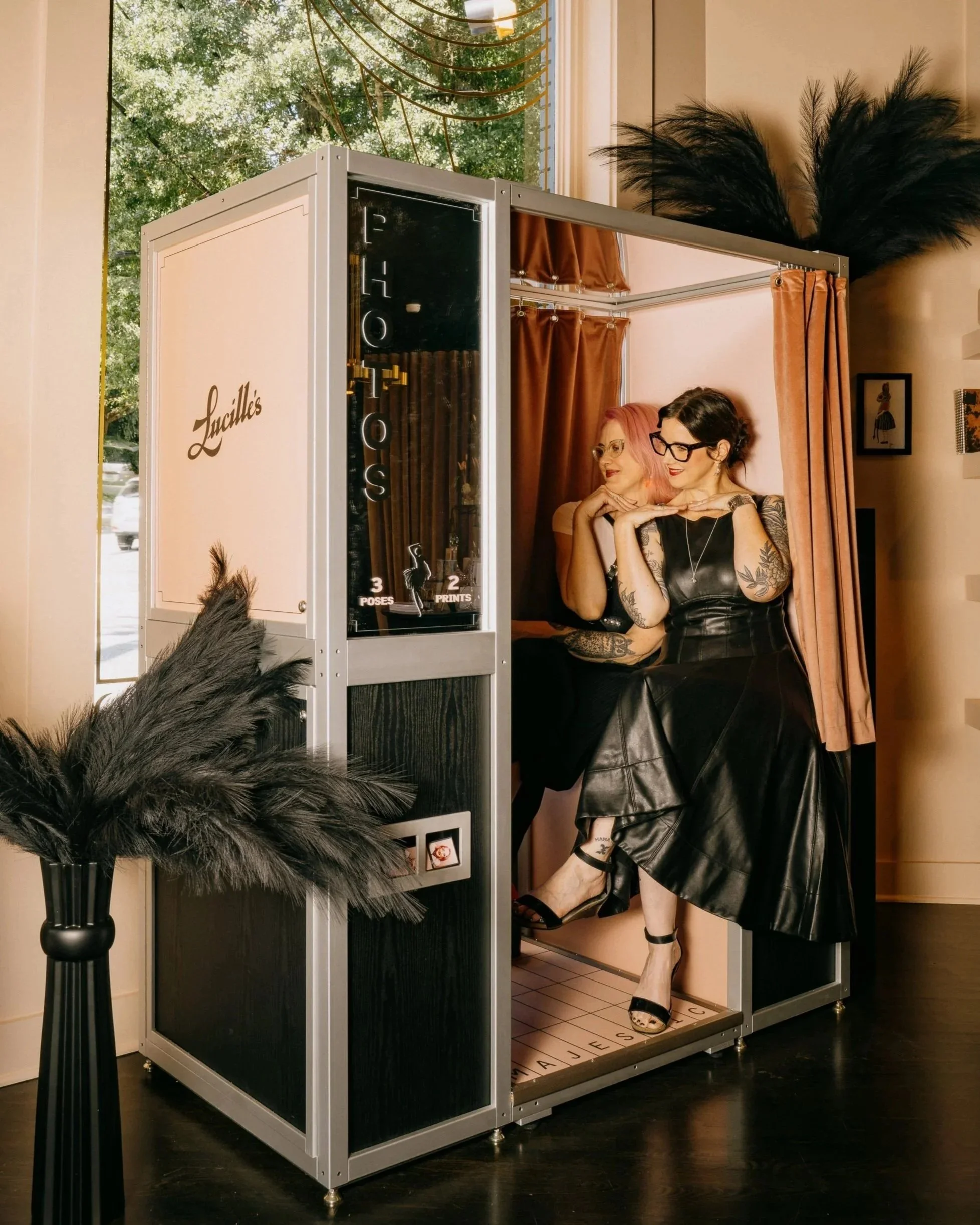 A photo booth with 'Lucille's' in elegant cursive font, set inside a cozy space. Two smiling women are seated inside the booth, one with pink hair and glasses, the other with dark hair and glasses. A tall black vase with black pampas grass.