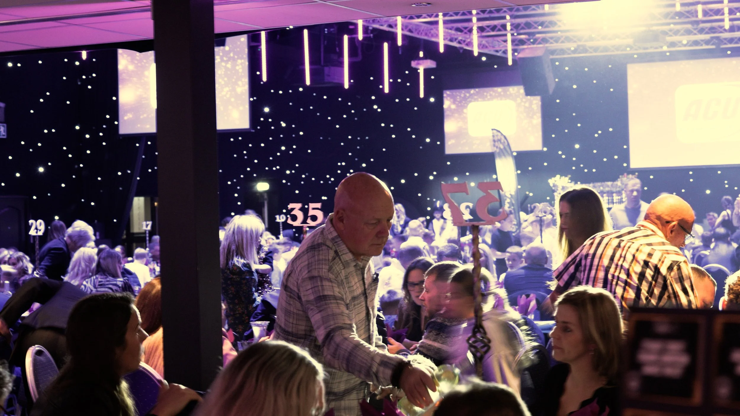 Crowd attending a large indoor event or conference with stage screens, some people standing and others sitting, and numbered signs for seating.