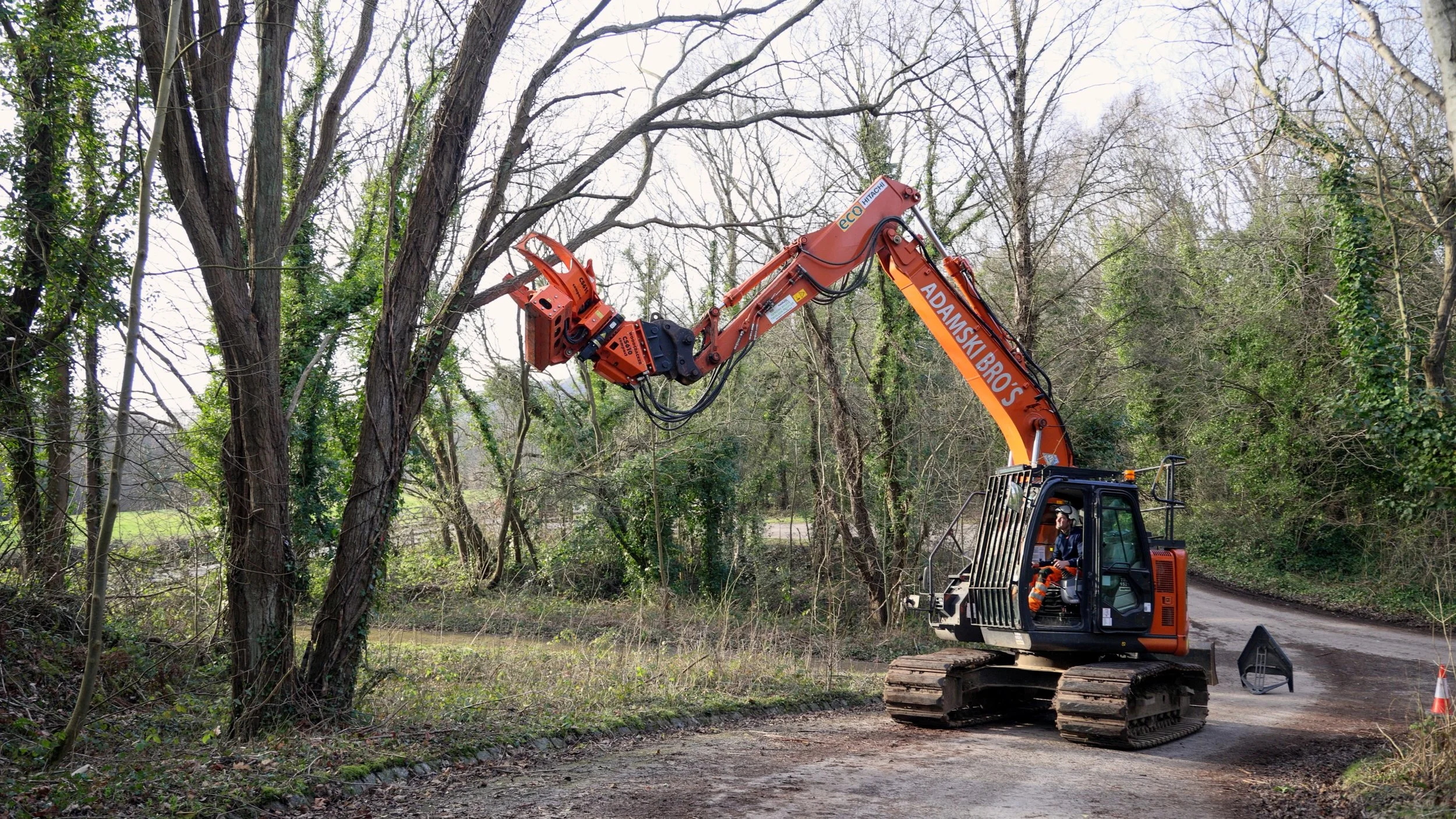A worker operating an orange excavator with the branding 'ADAMSKI BRO'S' on a dirt path in a wooded area with leafless trees and a grassy field in the background.