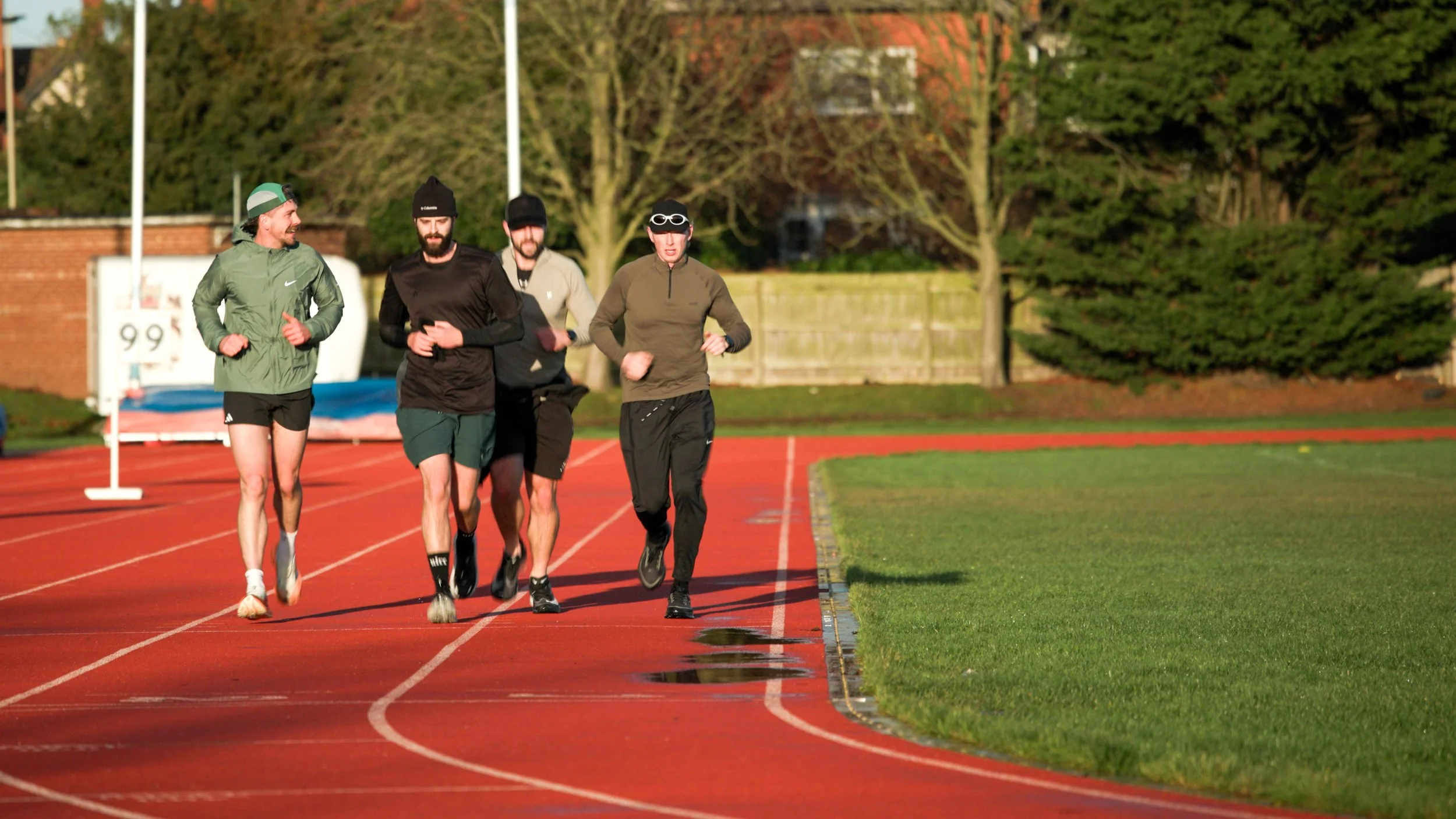 Four people running together on a red track, dressed in athletic clothing, with trees and a wooden fence in the background.