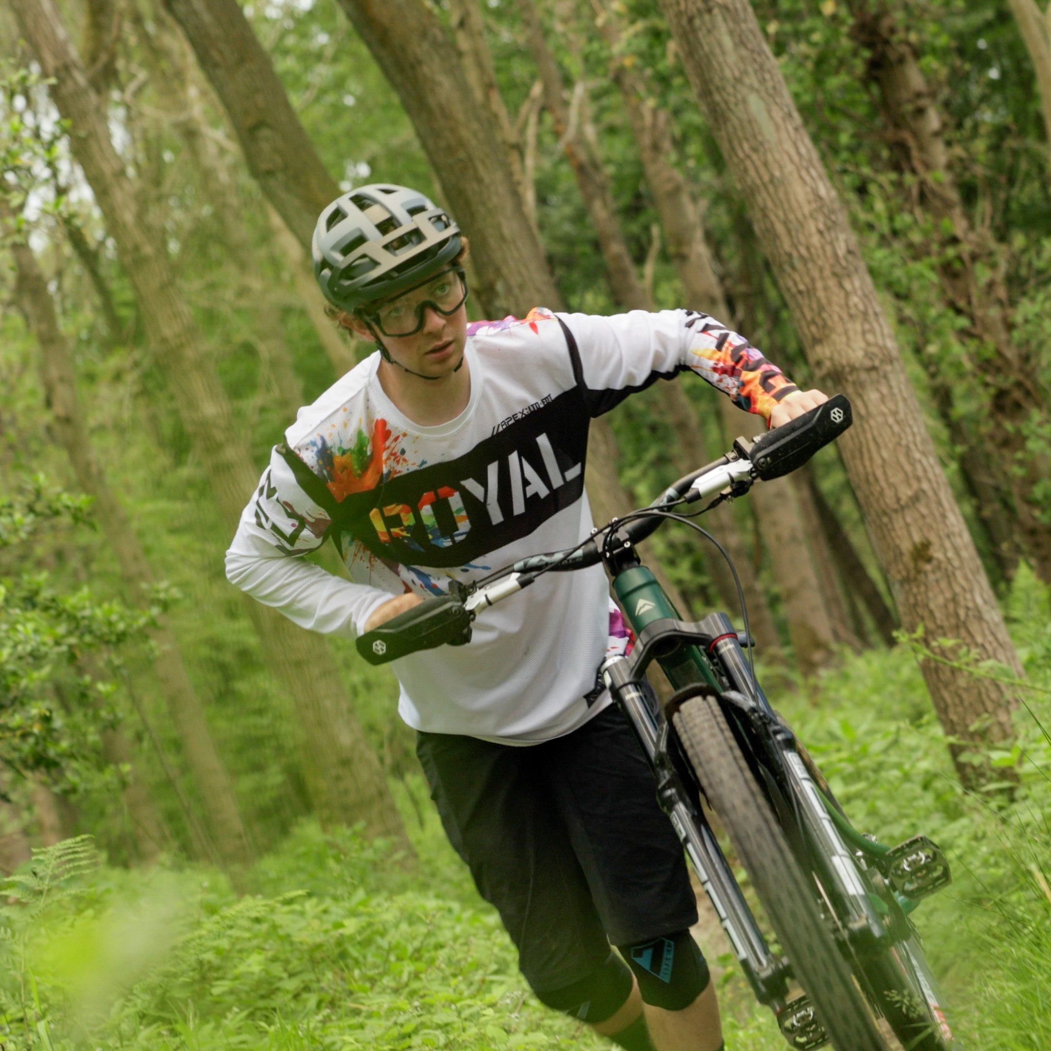 A man mountain biking on a trail in a green forest, wearing a helmet, glasses, and a colorful long-sleeve shirt.