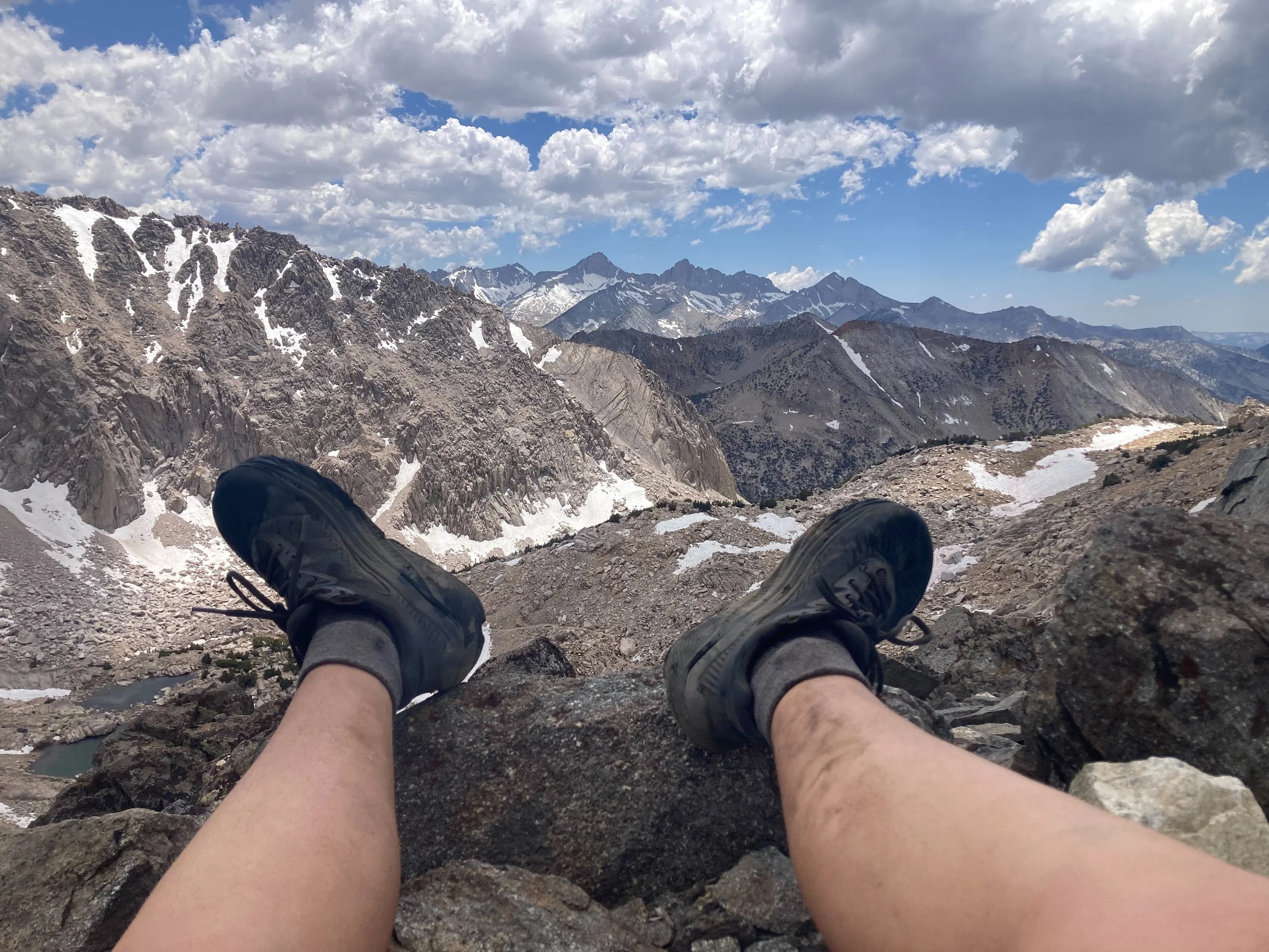 Person resting on a rocky mountain slope, overlooking a rugged mountain range with snow patches, under a partly cloudy sky.