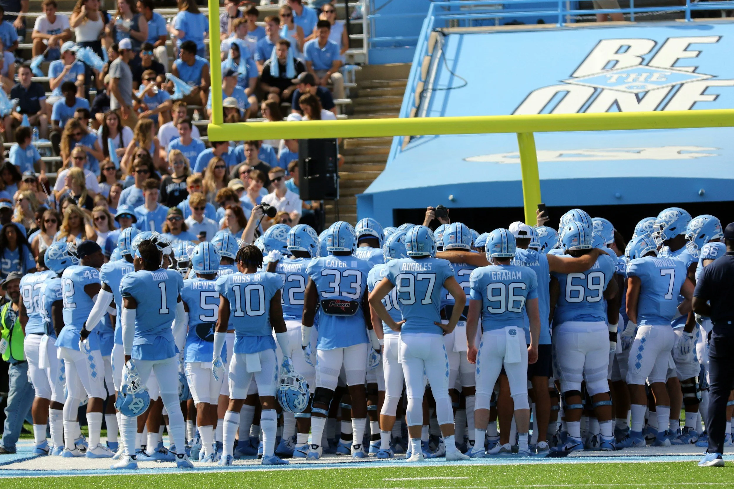 University of North Carolina football team standing on the field during a game, with fans seated in the stands behind them.