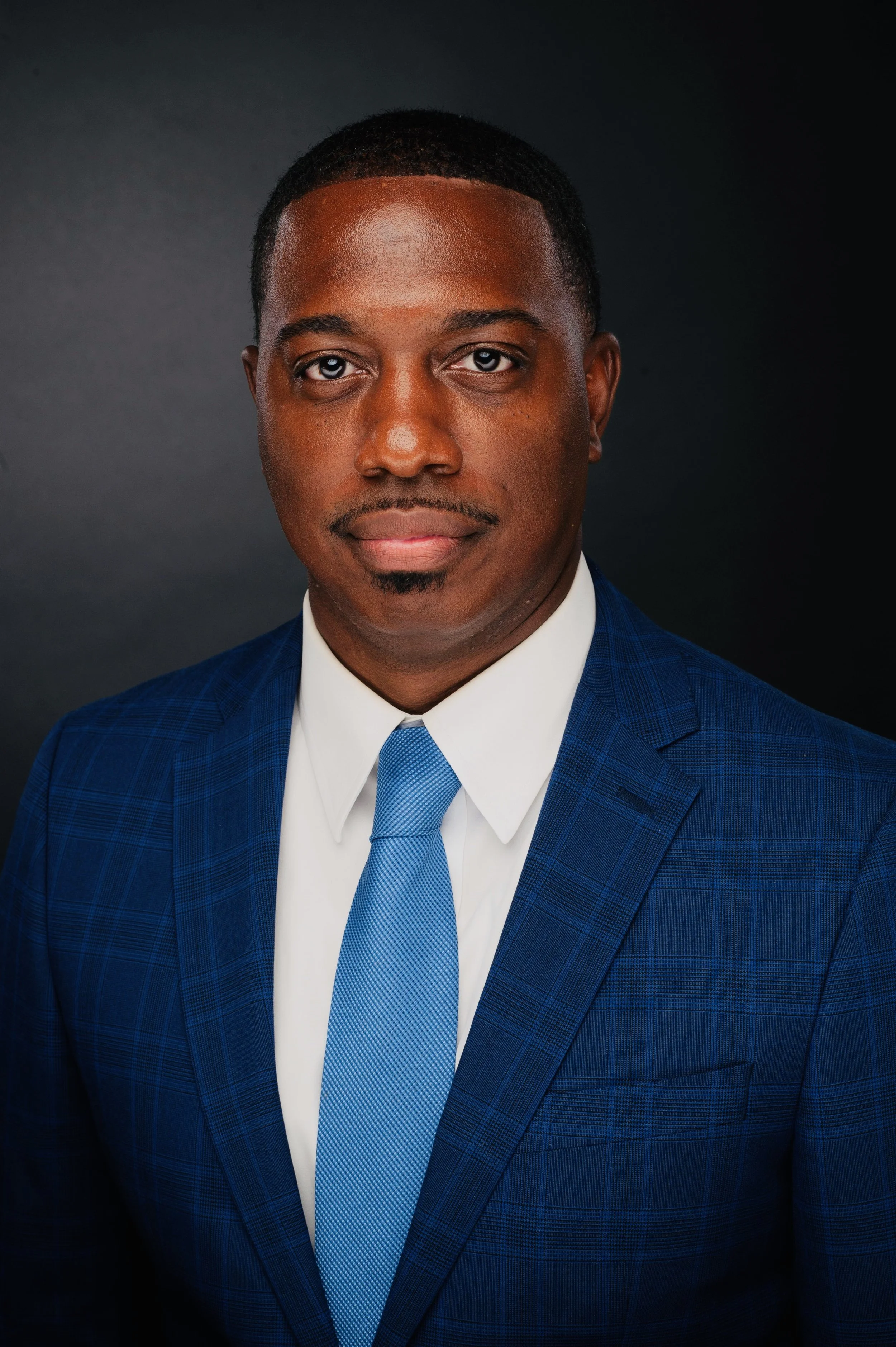 Professional headshot of an African American man wearing a blue suit, white shirt, and blue tie against a dark background.