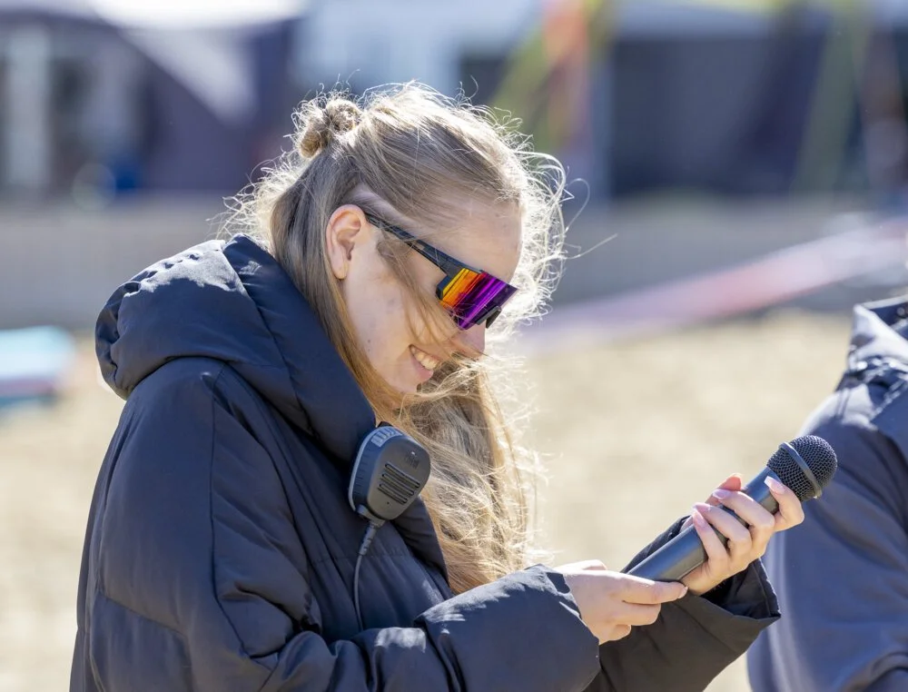 A young woman wearing rainbow-tinted sunglasses and a dark puffer jacket is holding and looking at a microphone while outdoors, smiling.