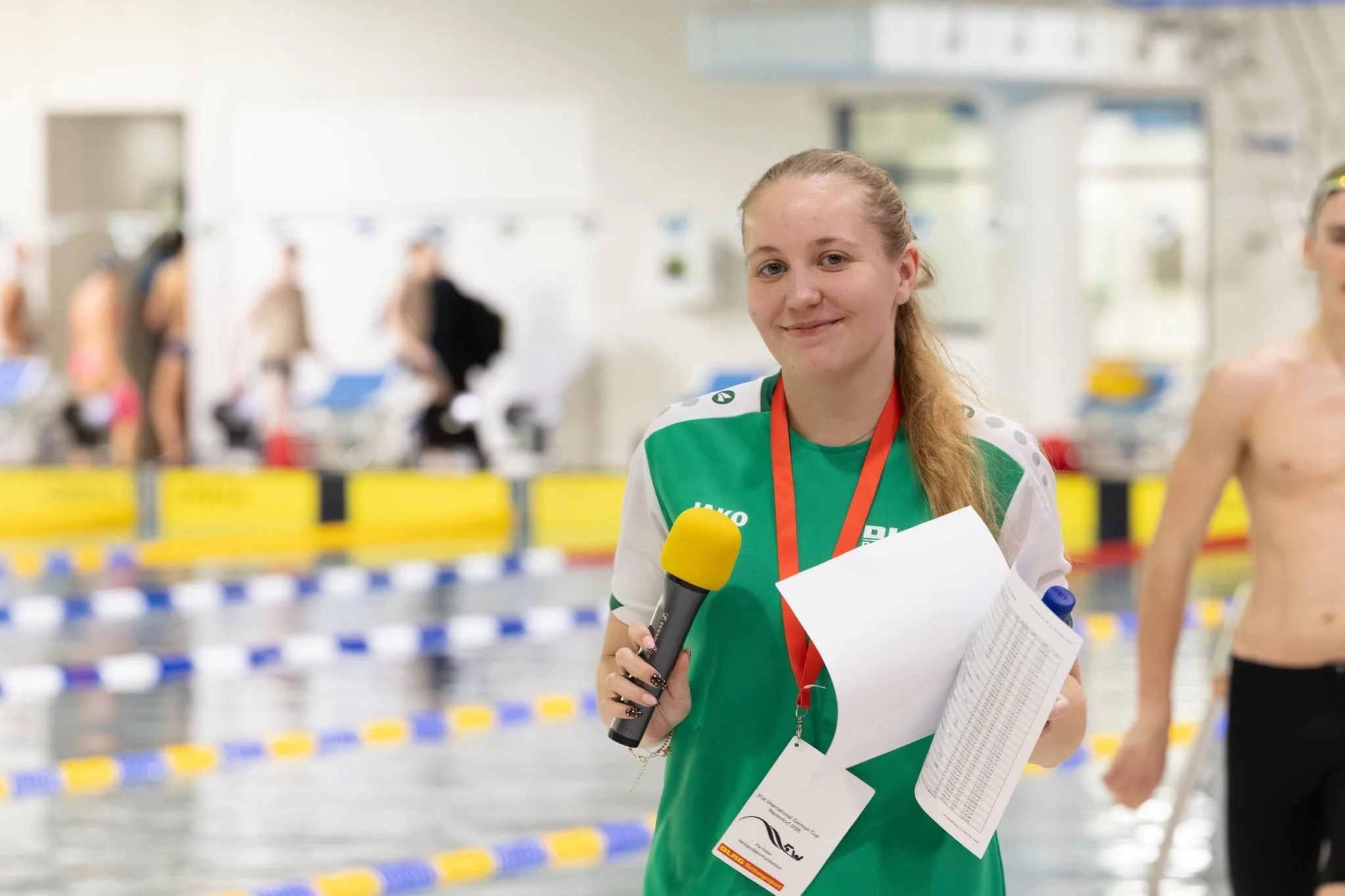 A young female swimmer with a medal around her neck, holding a microphone and papers, standing poolside at an indoor swimming competition with other swimmers and spectators in the background.