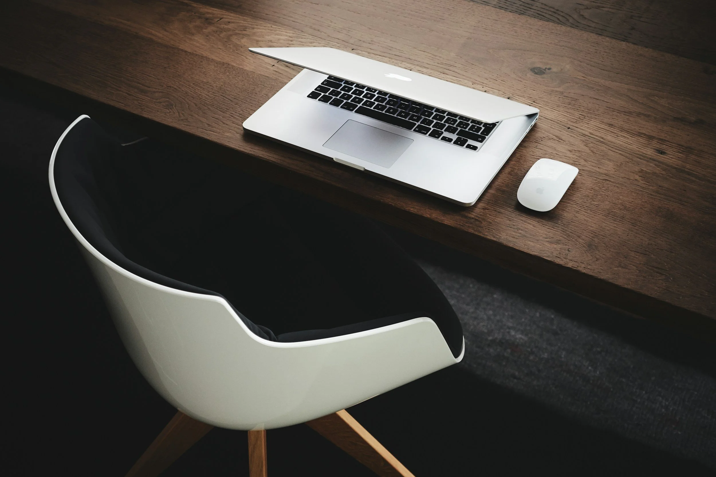 Un ordinateur portable blanc sur un bureau en bois, avec une souris blanche à côté, et une chaise noire partiellement visible.