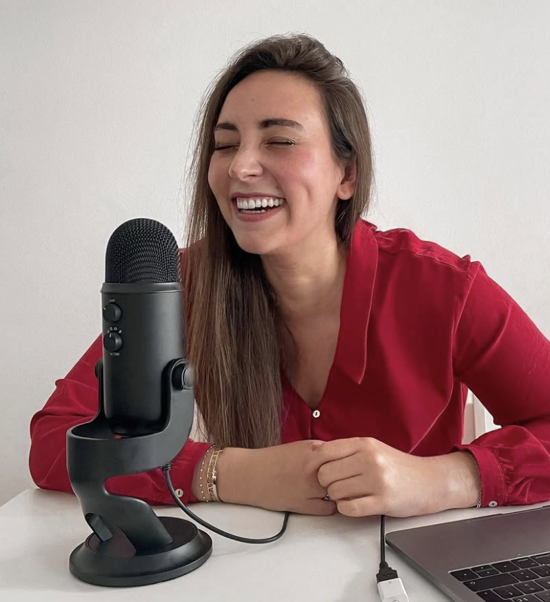 Une femme souriante avec une robe rouge, assise à un bureau avec un microphone, un ordinateur portable, et une ligne de câble.