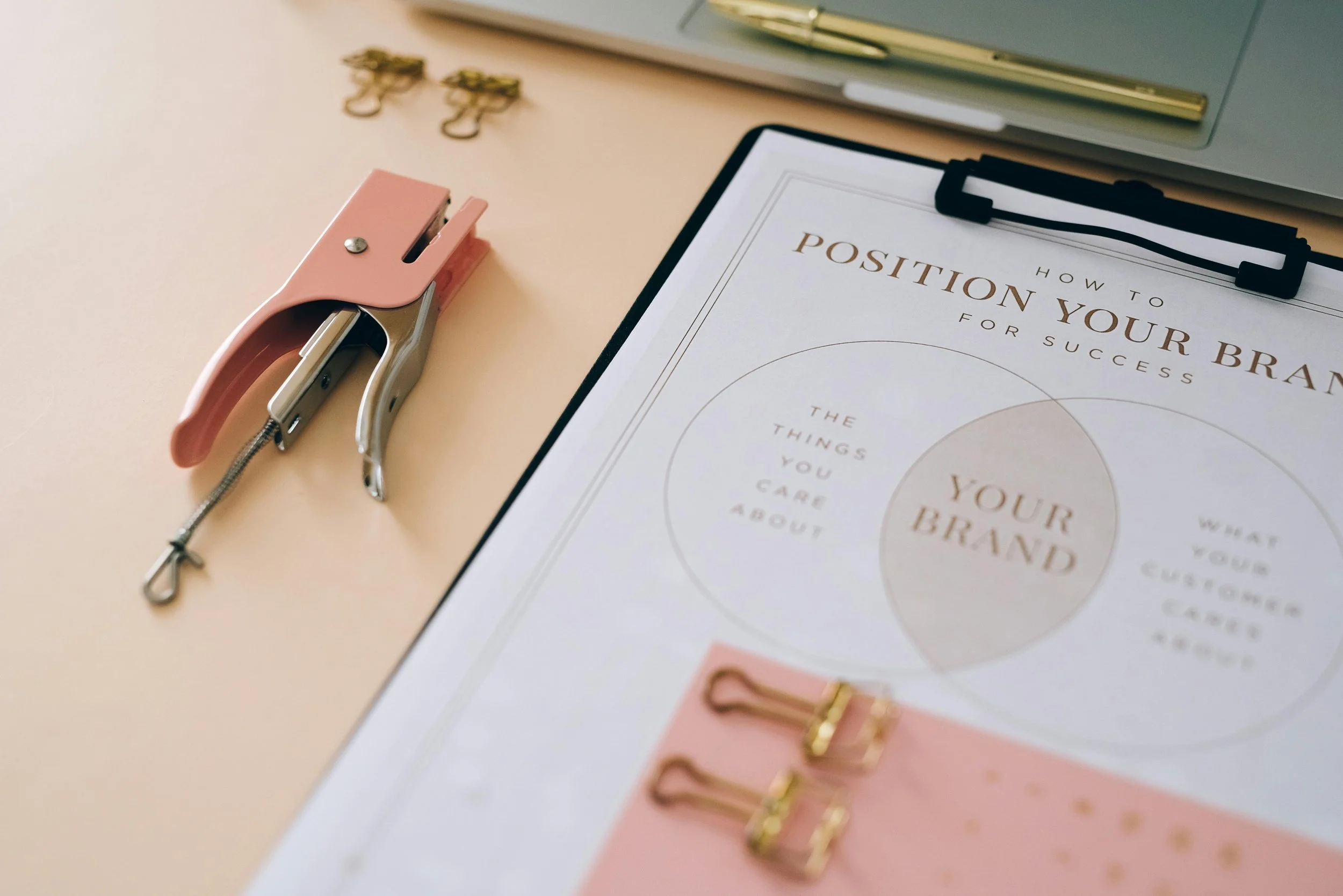 Une feuille de papier avec le titre « Comment valoriser votre marque » sur un classeur noir. Sur la table, il y a aussi un perforateur rose, deux pince-clip dorés, des agrafeuses, un carnet vert et un stylo.