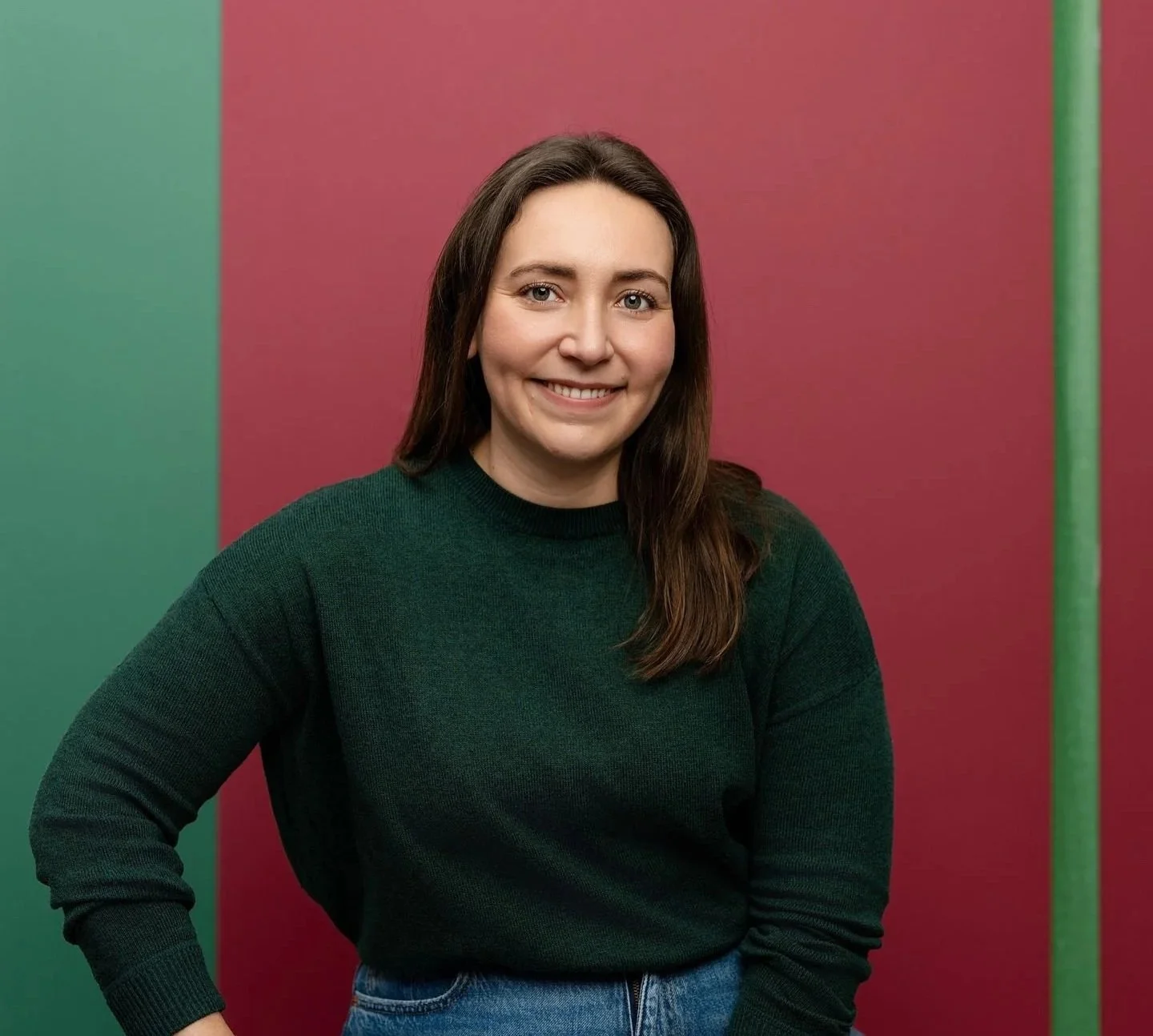 Une femme souriante avec des cheveux bruns, portant un pull vert foncé et un jean, pose devant un fond bicolore avec du vert à gauche et du rose à droite.